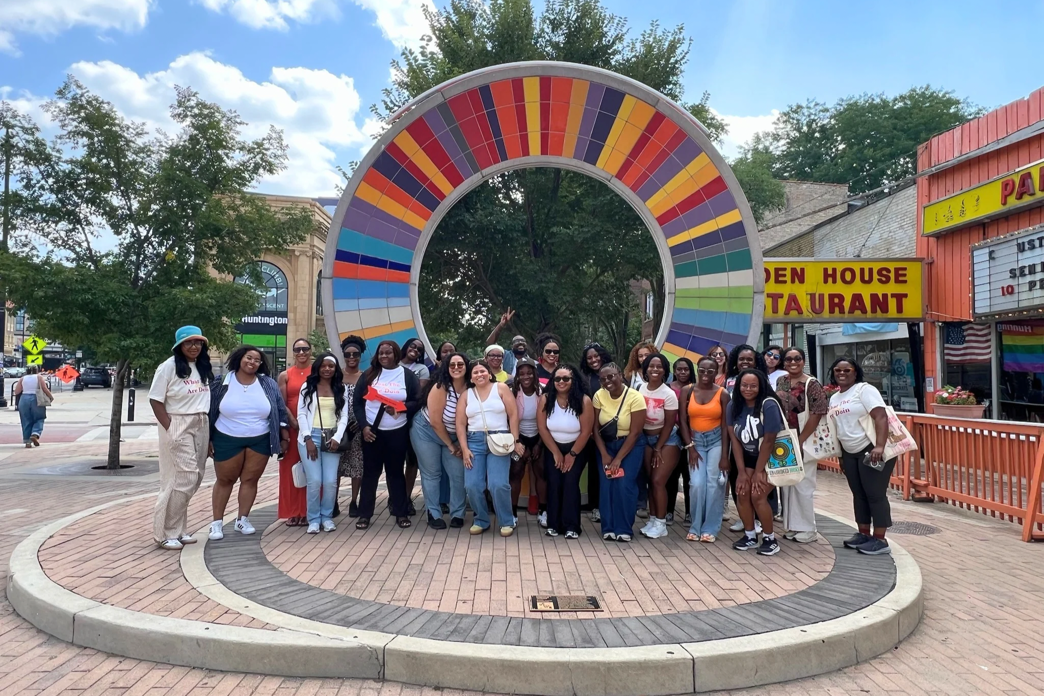 A large group of diverse people are posing together for a photo in front of a colorful circular art installation outdoors. There are trees, buildings, and a bright blue sky with clouds in the background.