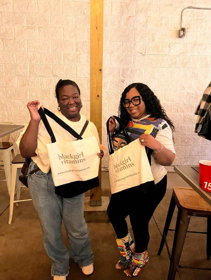 Two women smiling and holding tote bags with 'blackgirl vitamins' written on them, standing indoors against a brick wall.