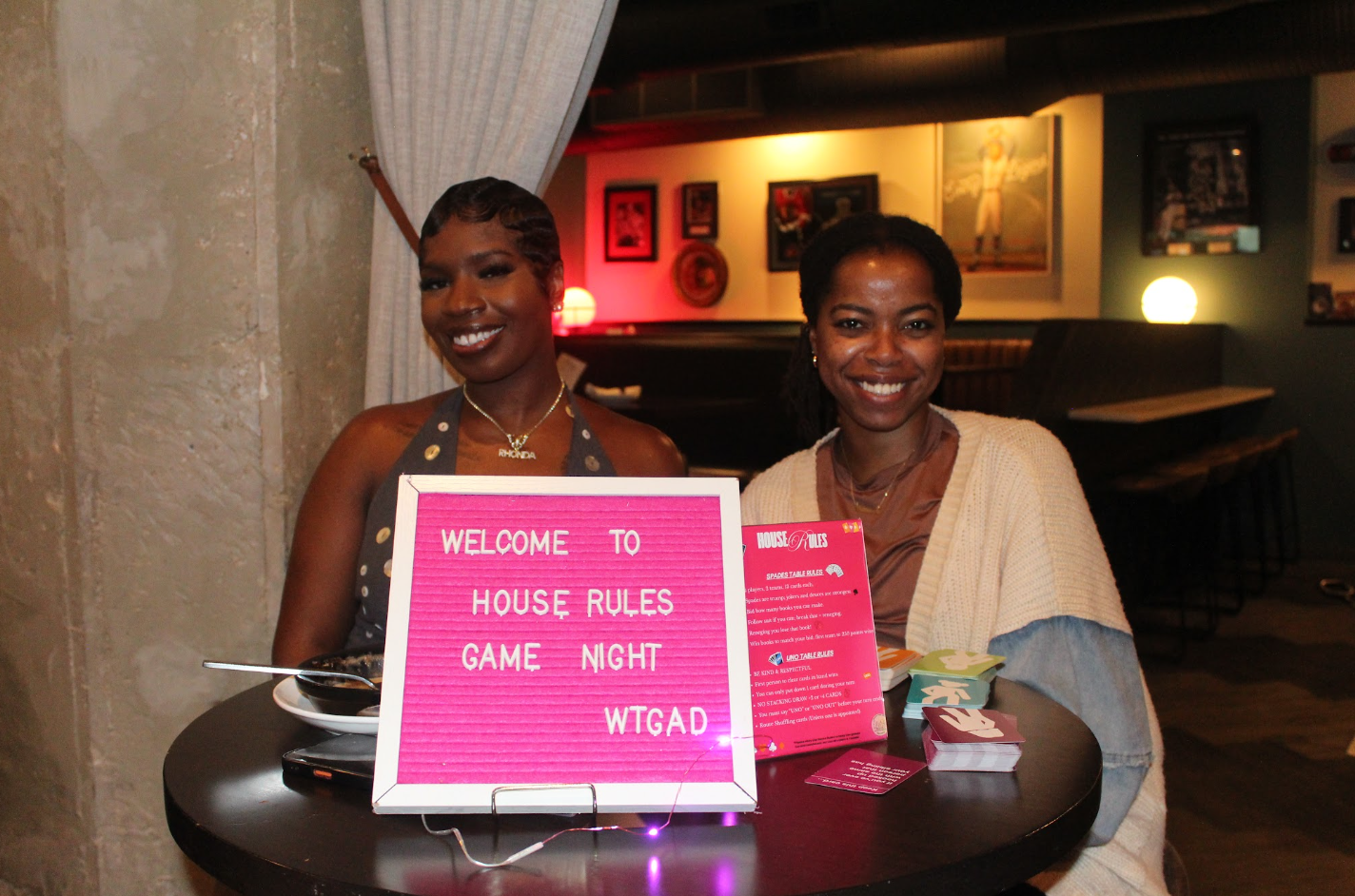 Two women are sitting at a round table in a dimly lit restaurant, smiling at the camera. One woman has short hair and is wearing a sleeveless top with a necklace, and the other woman has shoulder-length hair and is wearing a cream-colored sweater ove