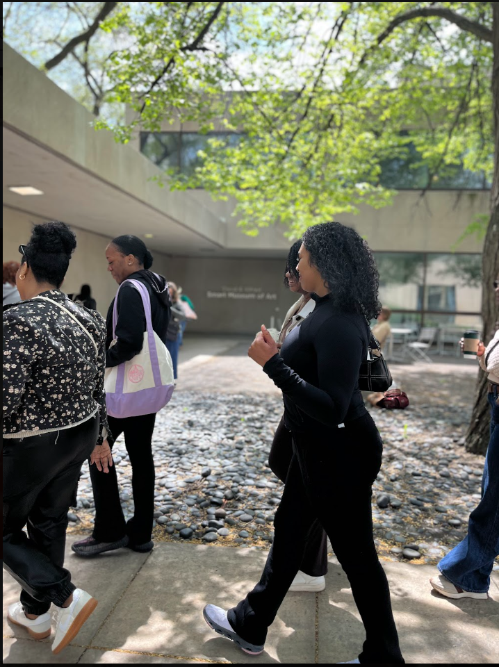 Group of people walking outdoors near a modern building with trees and sunlight overhead.