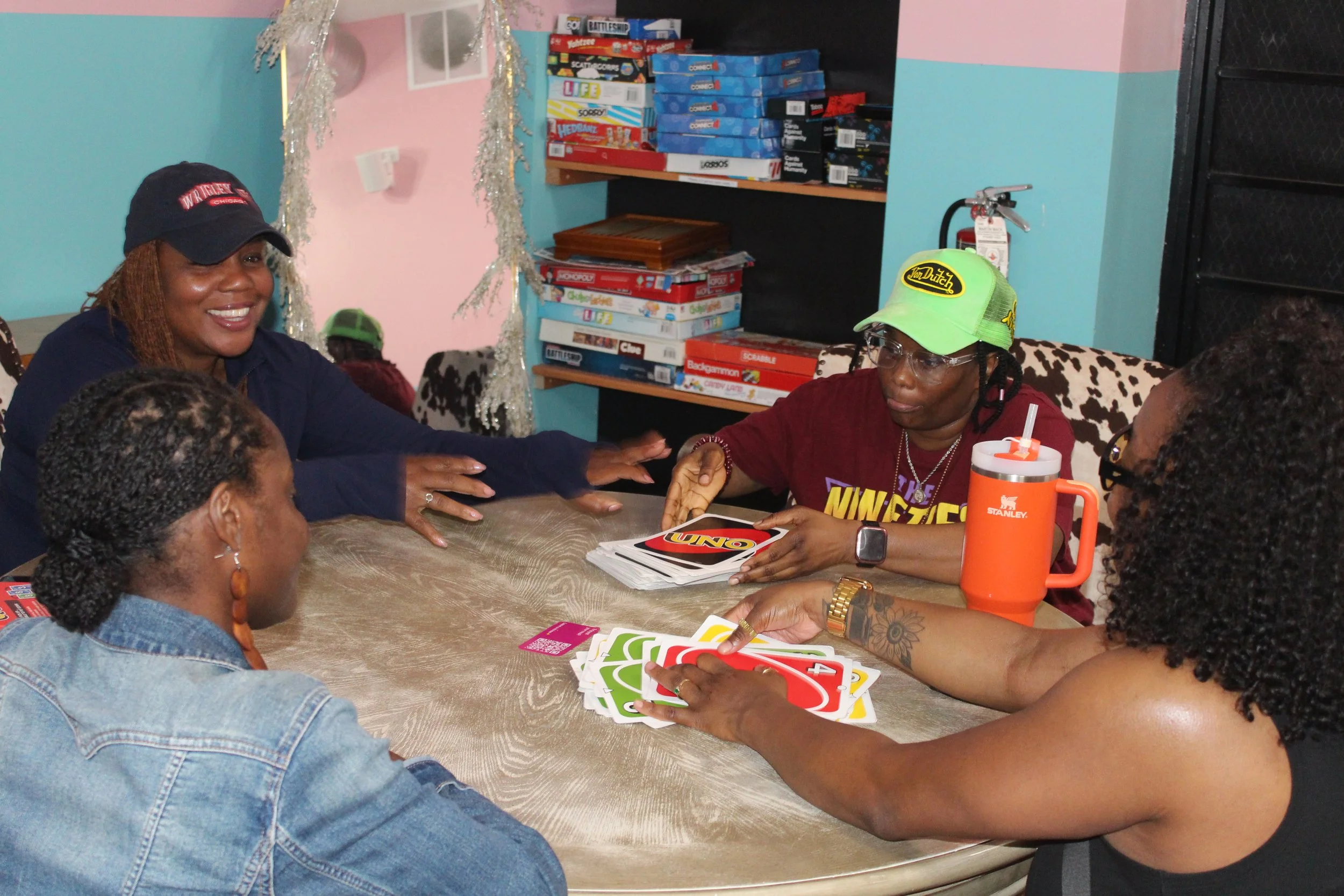 Group of women playing Uno card game at a table in a cozy room, with board games on shelves and colorful walls in the background.