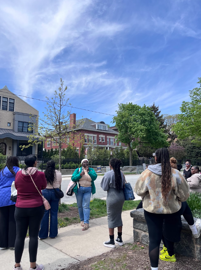 A group of diverse people gathered outdoors on a sidewalk, listening to a woman in a teal jacket who appears to be speaking. There are green trees, a red house and a blue sky with wispy clouds in the background.