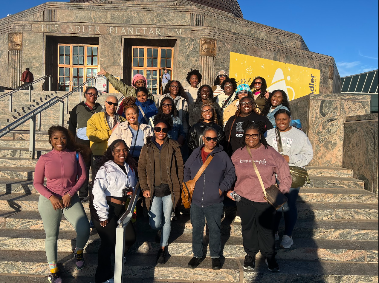Group of smiling women and girls posing on the stairs outside the Adler Planetarium in Chicago, with the building's entrance and a yellow banner for Adler Planetarium in the background.