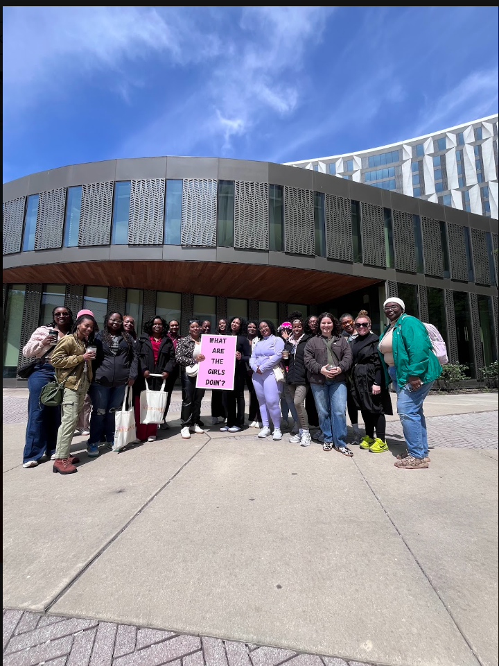 Group of women standing outdoors in front of a modern building, holding a pink sign that reads 'What are the girls doin?' sunny day with a blue sky.