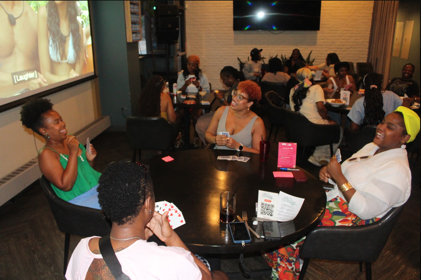 Group of women sitting around a black table in a restaurant, playing cards and laughing, with a TV on the wall displaying a video.