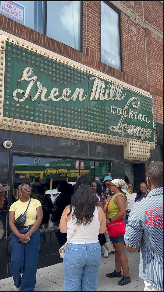 A group of people standing outside the Green Mill Cooktail Lounge, a bar or lounge, with a vintage marquee sign on a brick building. The sign has the name 'Green Mill' and other decorative elements. The scene shows a diverse crowd, some wearing casua