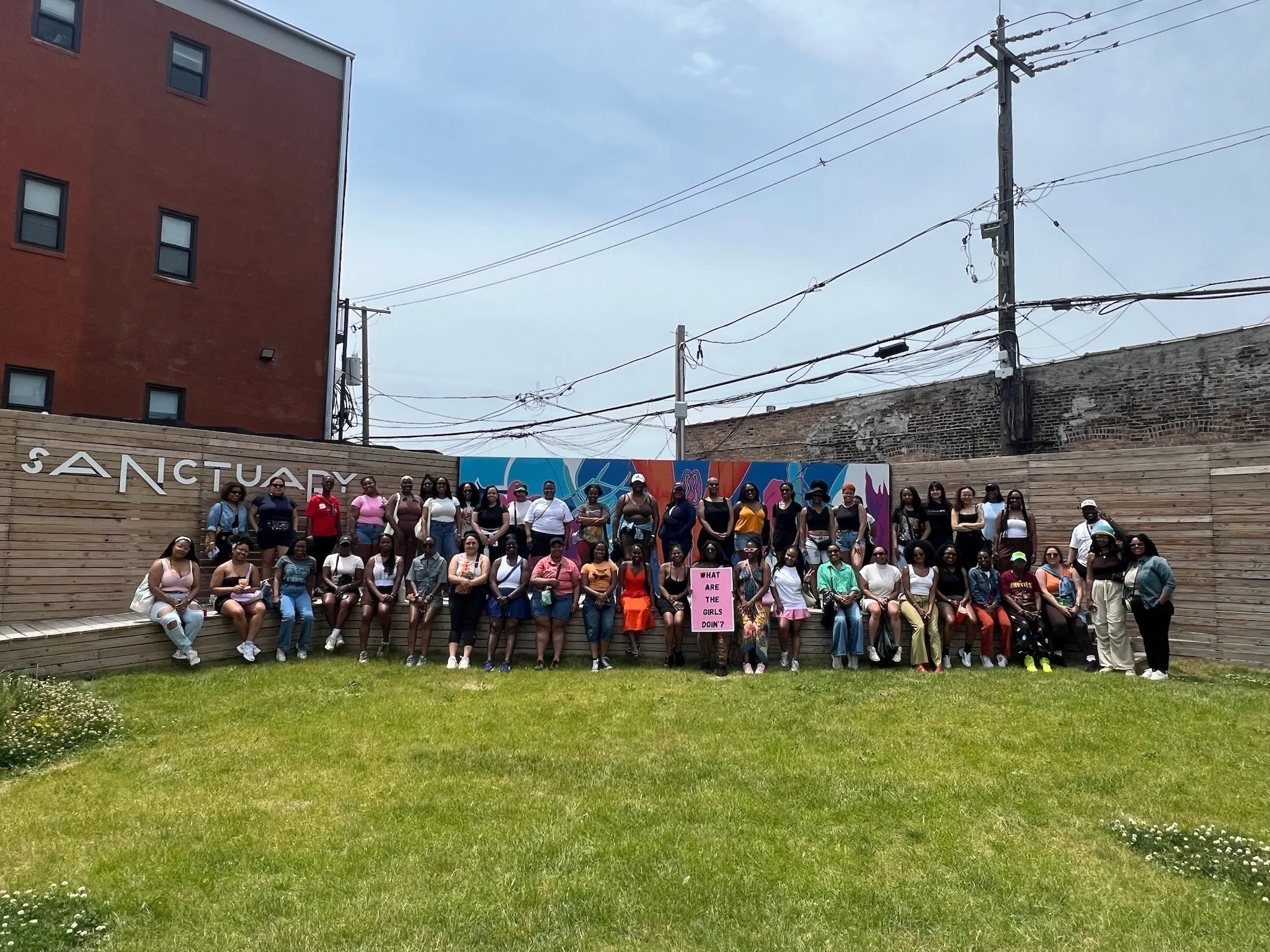 Group of women standing on a wooden platform outdoors in front of a colorful mural with the word 'Sanctuary' and holding a pink sign that reads 'What are the girls doing?'. Buildings, power lines, and a clear sky are in the background.