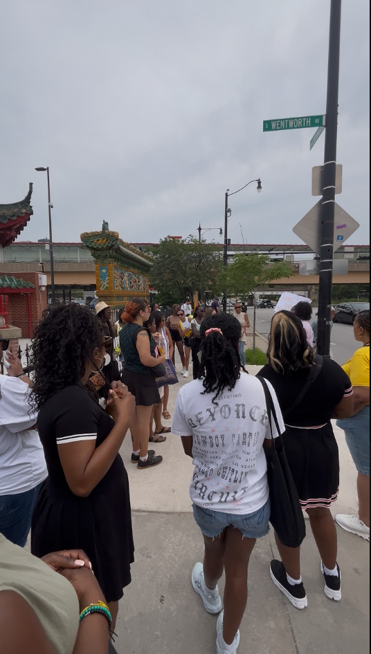 A diverse group of people standing on a sidewalk in an urban area, with a street sign reading 'S Wentworth W' visible. Some individuals are wearing masks, and there are traditional Asian-style decorative elements in the background, along with trees a