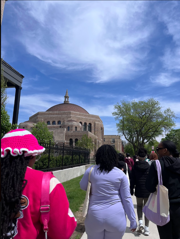 Group of diverse people walking on a sidewalk toward a large church or cathedral with a dome and a small spire, surrounded by trees, under a blue sky with clouds.