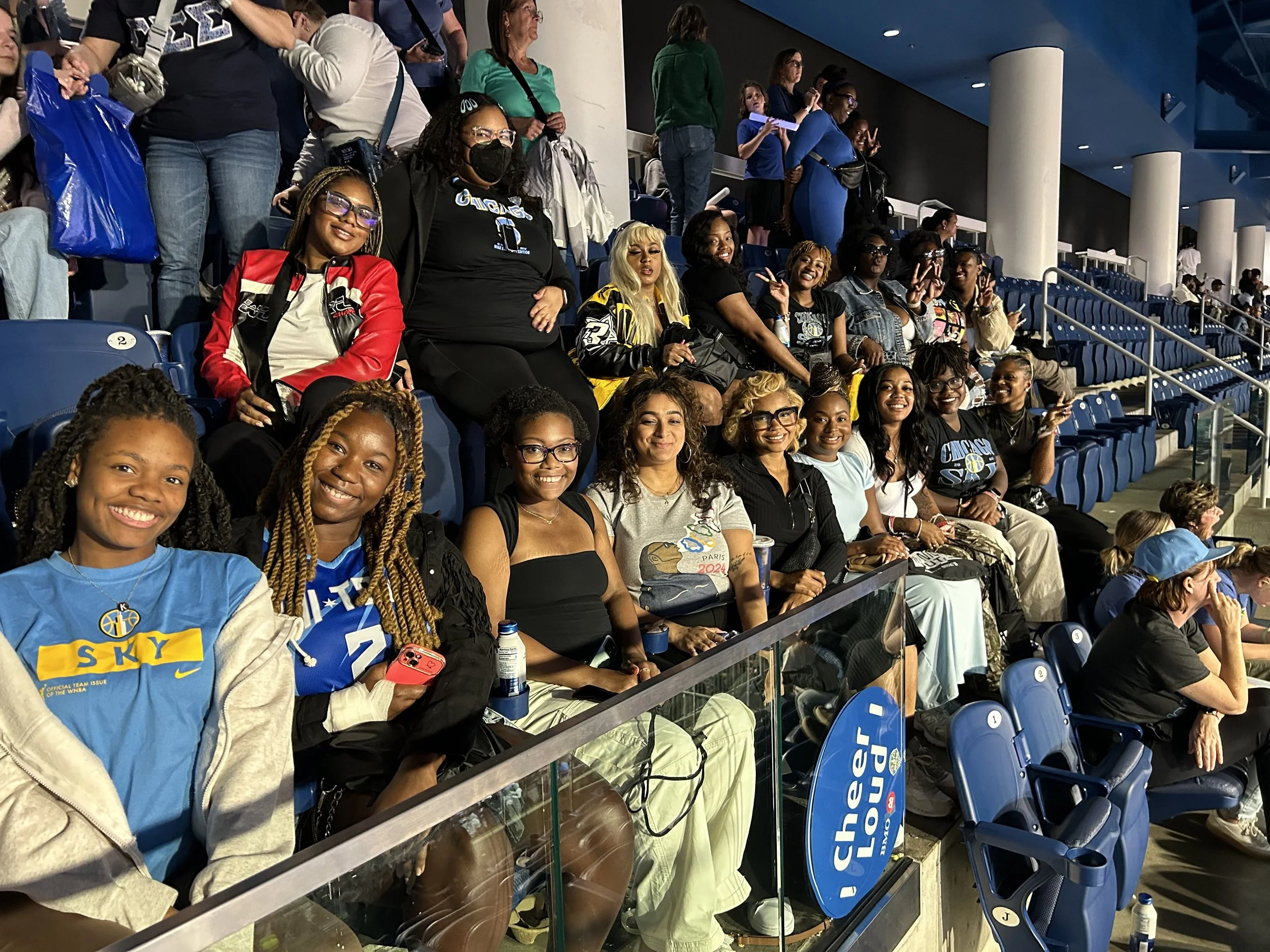 Group of women and girls sitting and standing in a stadium seating area, smiling, some making peace signs, with some wearing blue and black shirts, surrounded by empty blue seats and a glass barrier.