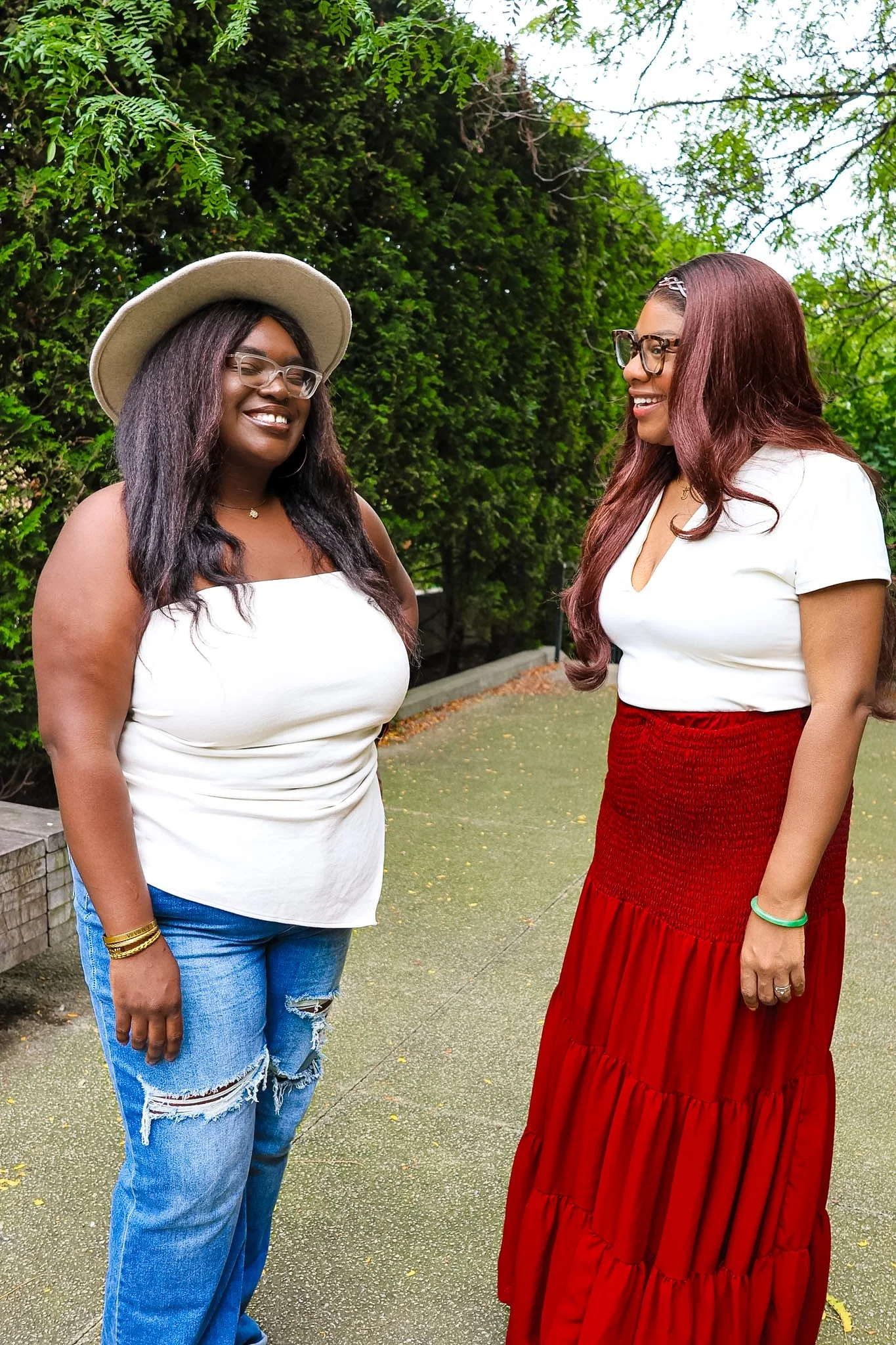 Two women smiling and talking outdoors, surrounded by greenery. One woman is wearing a white sleeveless top, distressed jeans, and a hat, while the other is wearing glasses, a white top, and a long red skirt.