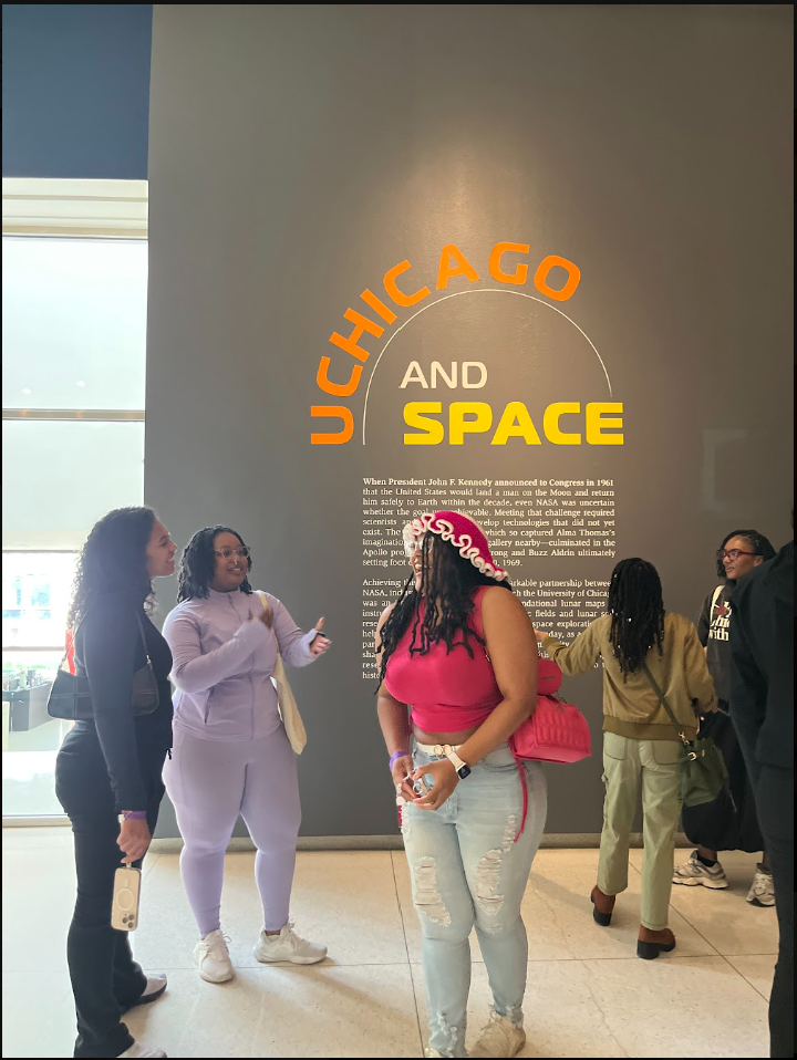 Group of five women in casual clothing standing in front of a gray wall with a large text display reading 'Chicago and Space' and additional informational text about NASA and space exploration.