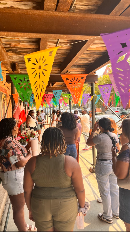 People gathered under a wooden overhang decorated with colorful paper banners for a celebration or event on a sunny day.