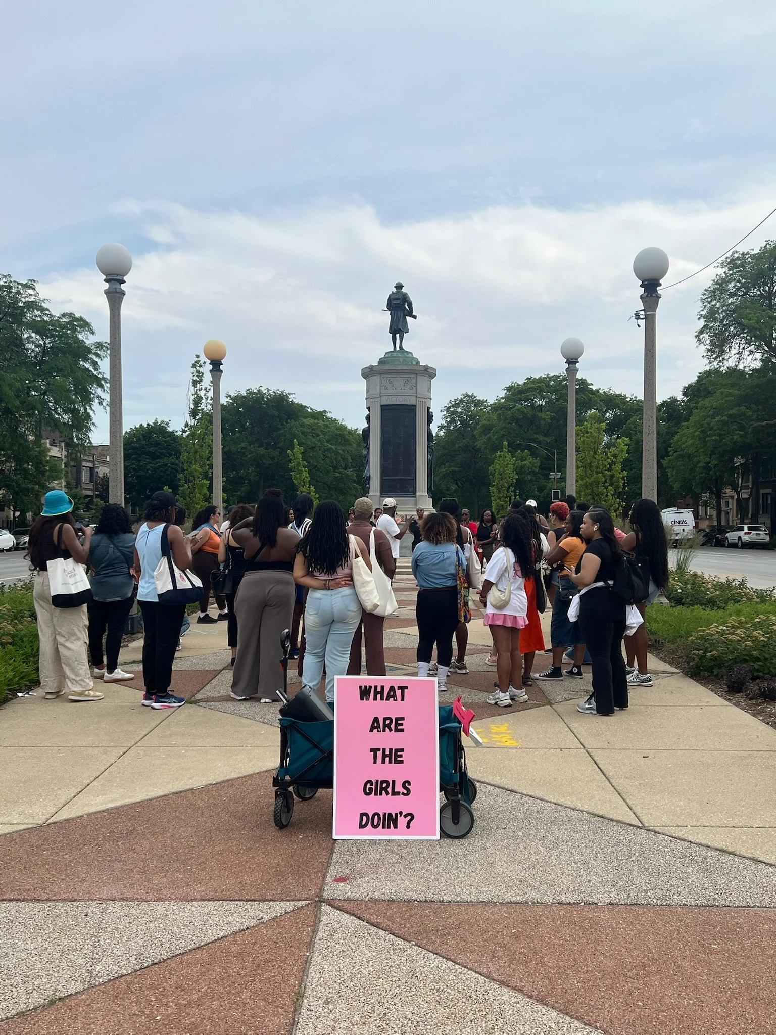 A group of women and girls gathered in a city square around a statue of a historical figure standing on a tall pedestal. In the foreground, a pink sign reads, 'What are the girls doin'?' and the crowd appears to be listening or participating in an ev