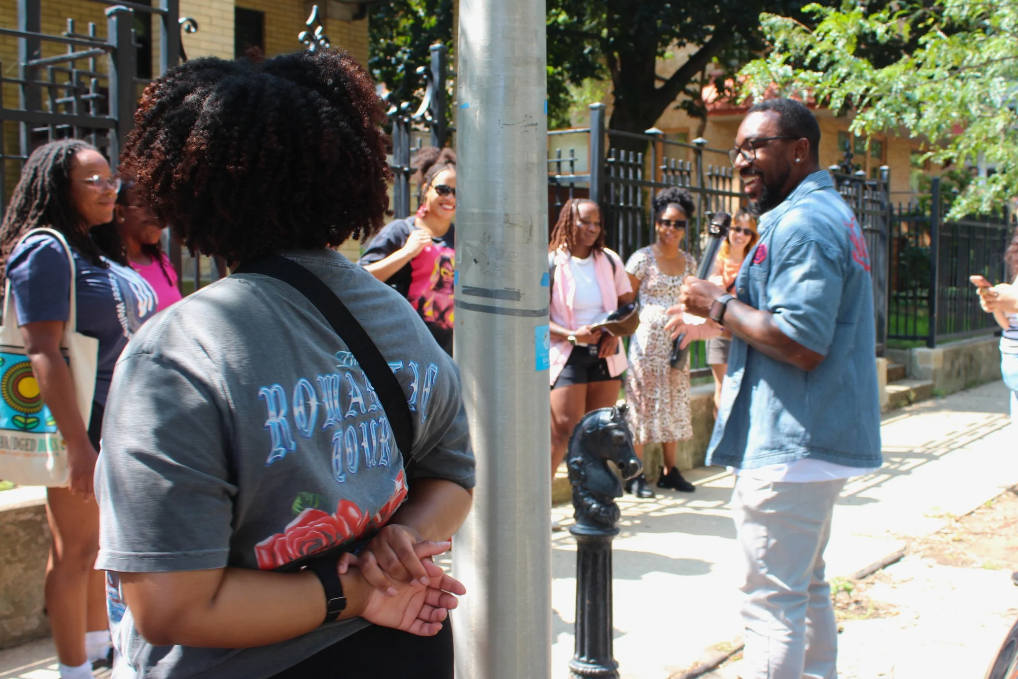 A group of people including a man holding a microphone and several women, engaging in conversation outdoors on a sunny day near a street and fence.