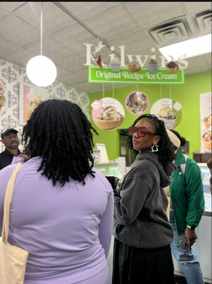 Women standing in line inside an ice cream shop, with a sign that says 'Livins Original Recipe Ice Cream' hanging above. The shop has a bright green wall and menu boards with ice cream images.