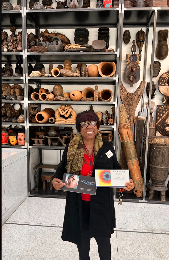 A woman standing in front of a display of various African art and artifacts, holding a certificate and a booklet, smiling at the camera.