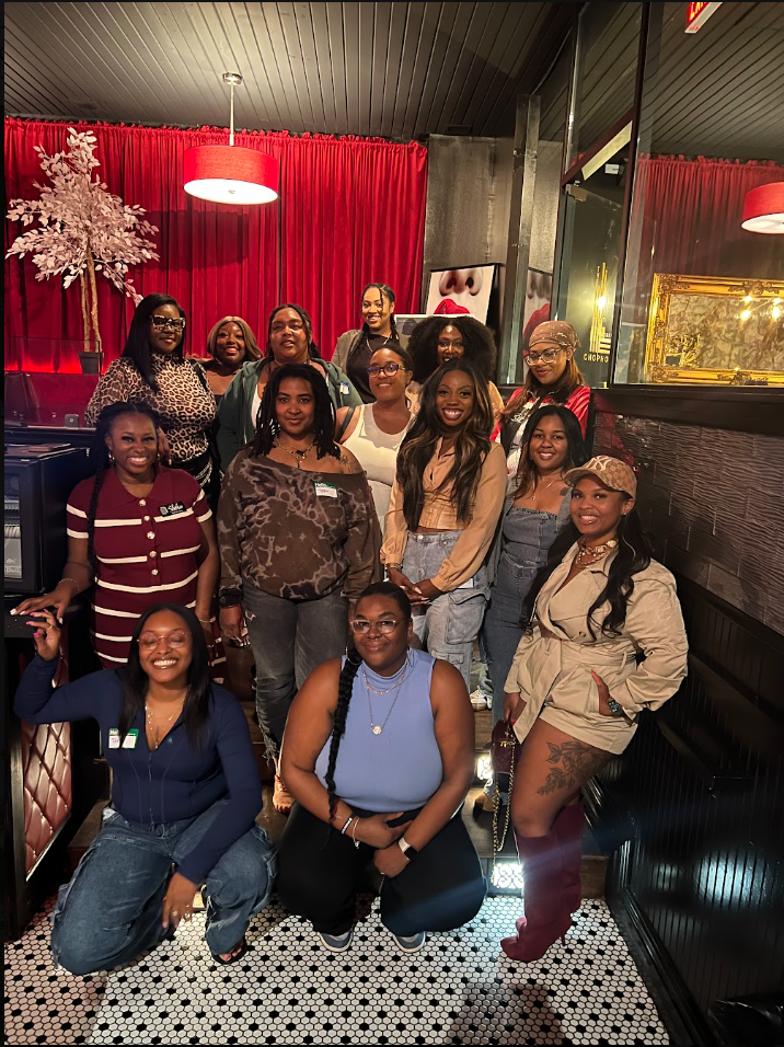 Group of women posing together in a decorated restaurant with red curtains and artwork, some with glasses, and one with a hat.