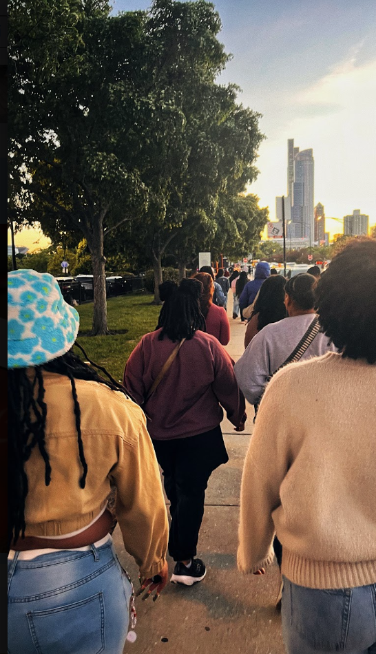 People walking in a line along a city sidewalk during sunset, with green trees on the left and a skyline with tall buildings in the background.