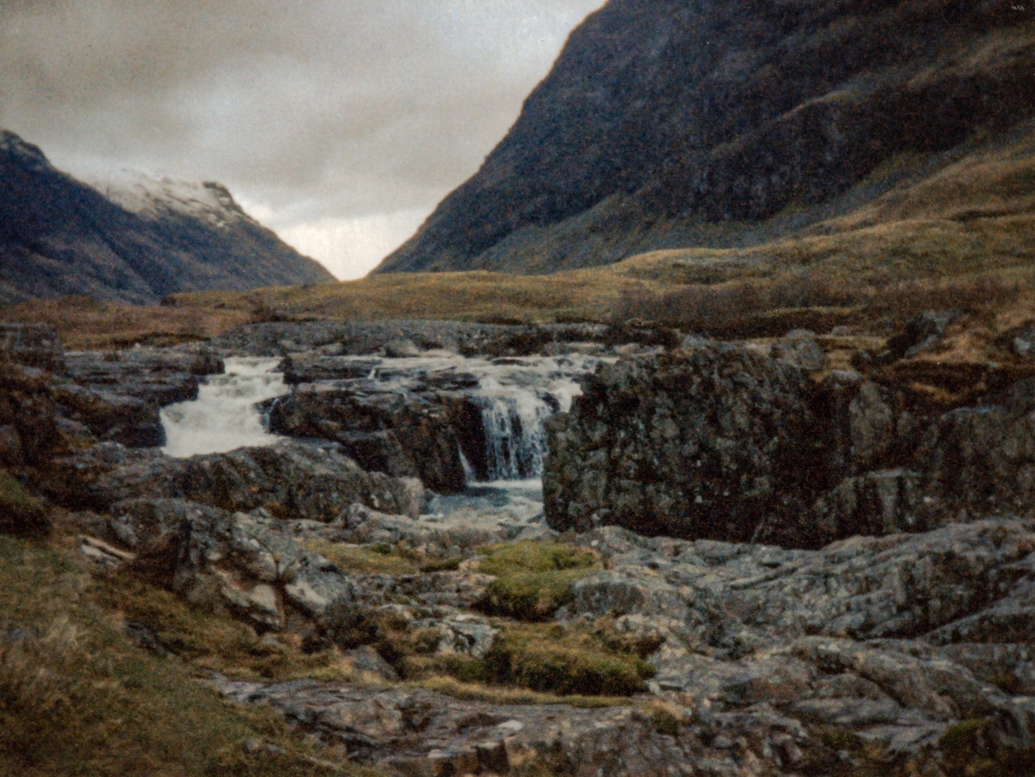 A rugged mountain landscape with steep dark rocky slopes, a small waterfall in the foreground, and cloudy sky overhead.