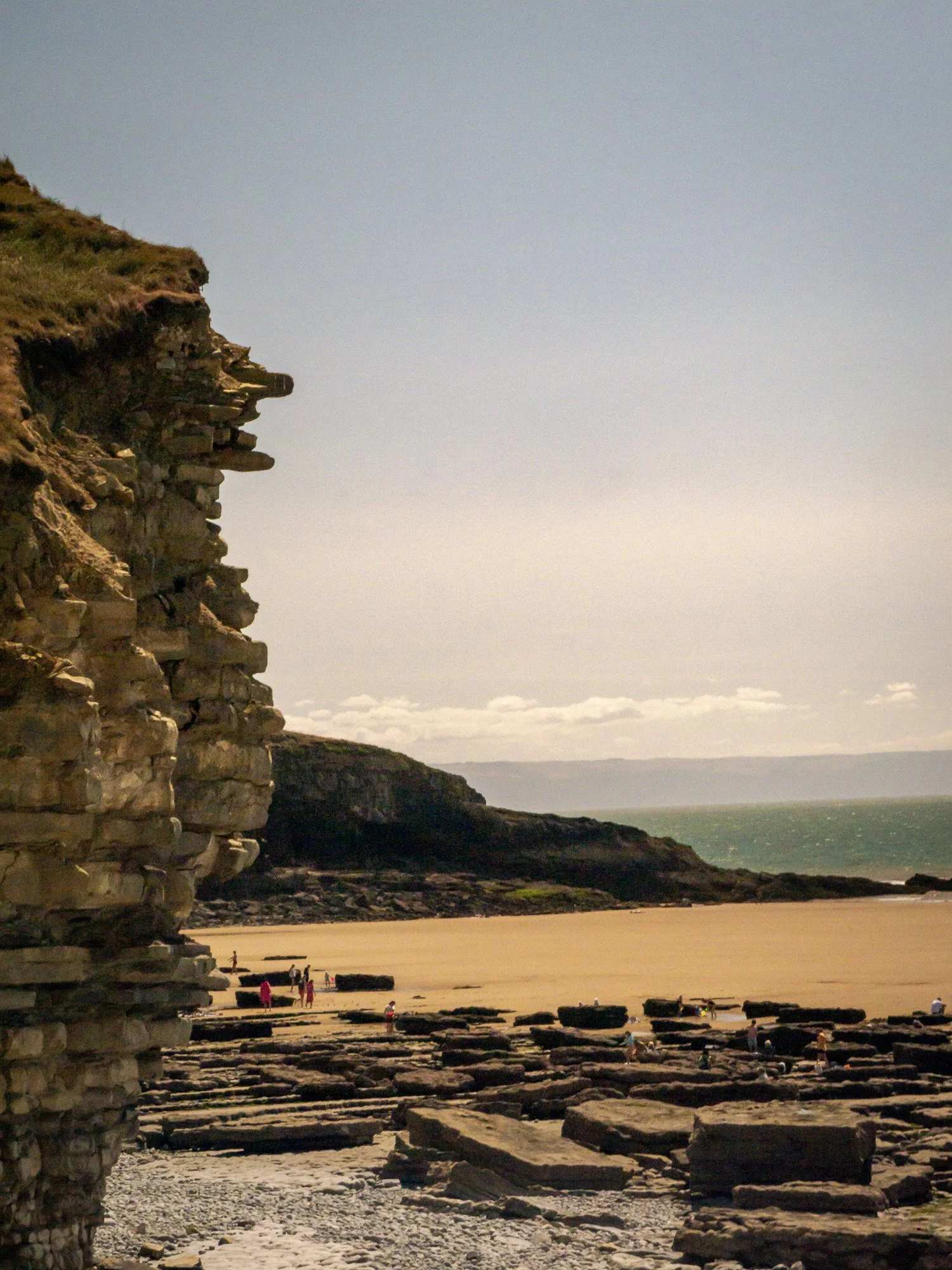 Cliffs on the coast overlooking the ocean with people walking along the beach and rock formations in the foreground.