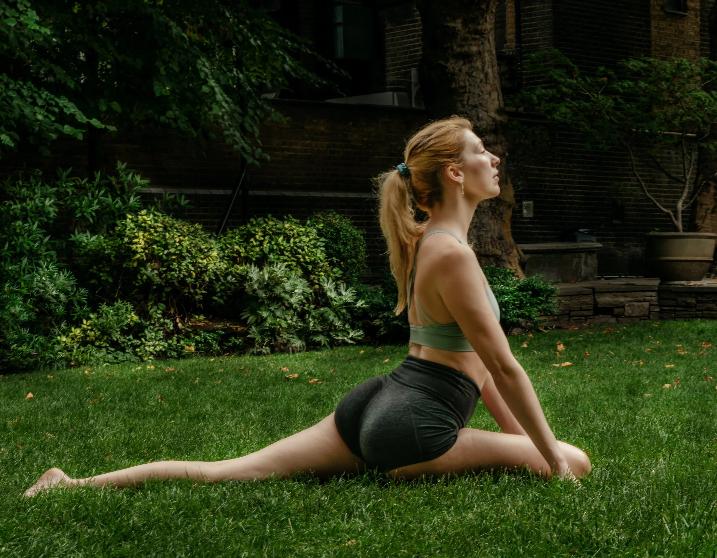 A woman practicing yoga outdoors on a grassy area near trees and bushes, with her hair tied back in a ponytail, wearing a tank top and shorts, performing a lunge with her right leg forward and left leg extended back.