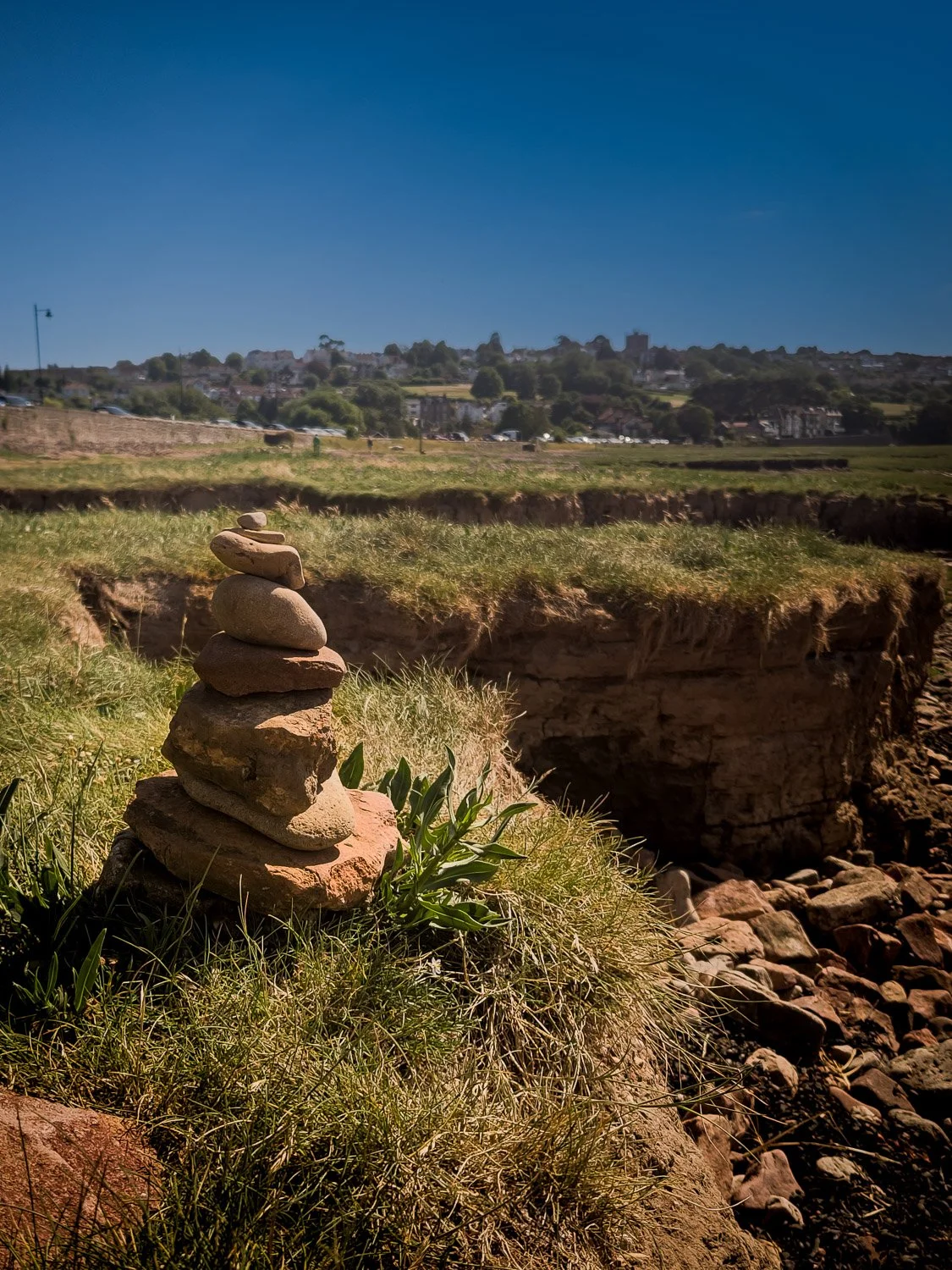 Stacked rocks balancing on grass near a cliff edge with a cityscape in the background under a blue sky.
