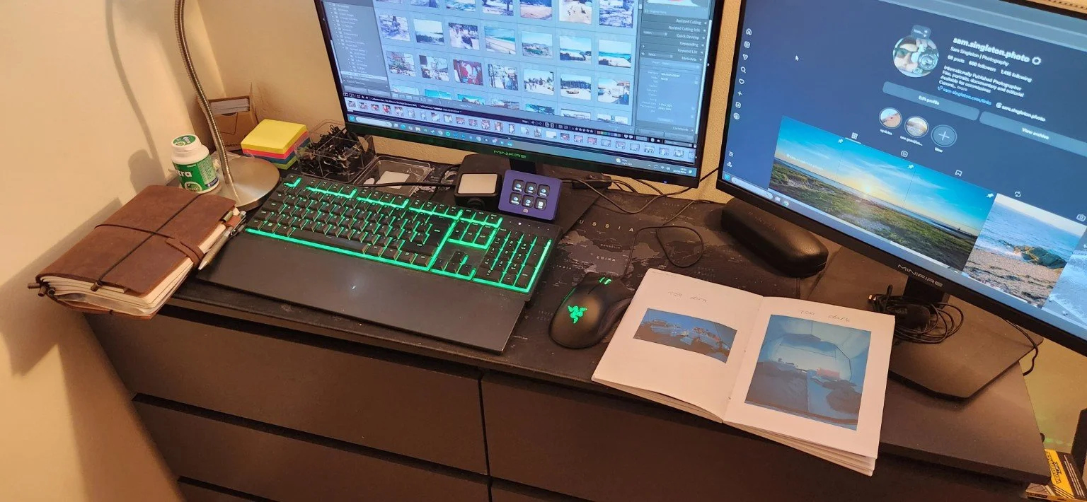 A dual-monitor computer setup on a dark desk with a graphics editing program on the left monitor and a social media profile on the right monitor. The desk has a brown leather-bound notebook, a small container of supplements, a desk lamp, sticky notes, a small tray with miscellaneous items, a keyboard with green backlighting, a gaming mouse, and an open photo album with landscape pictures.