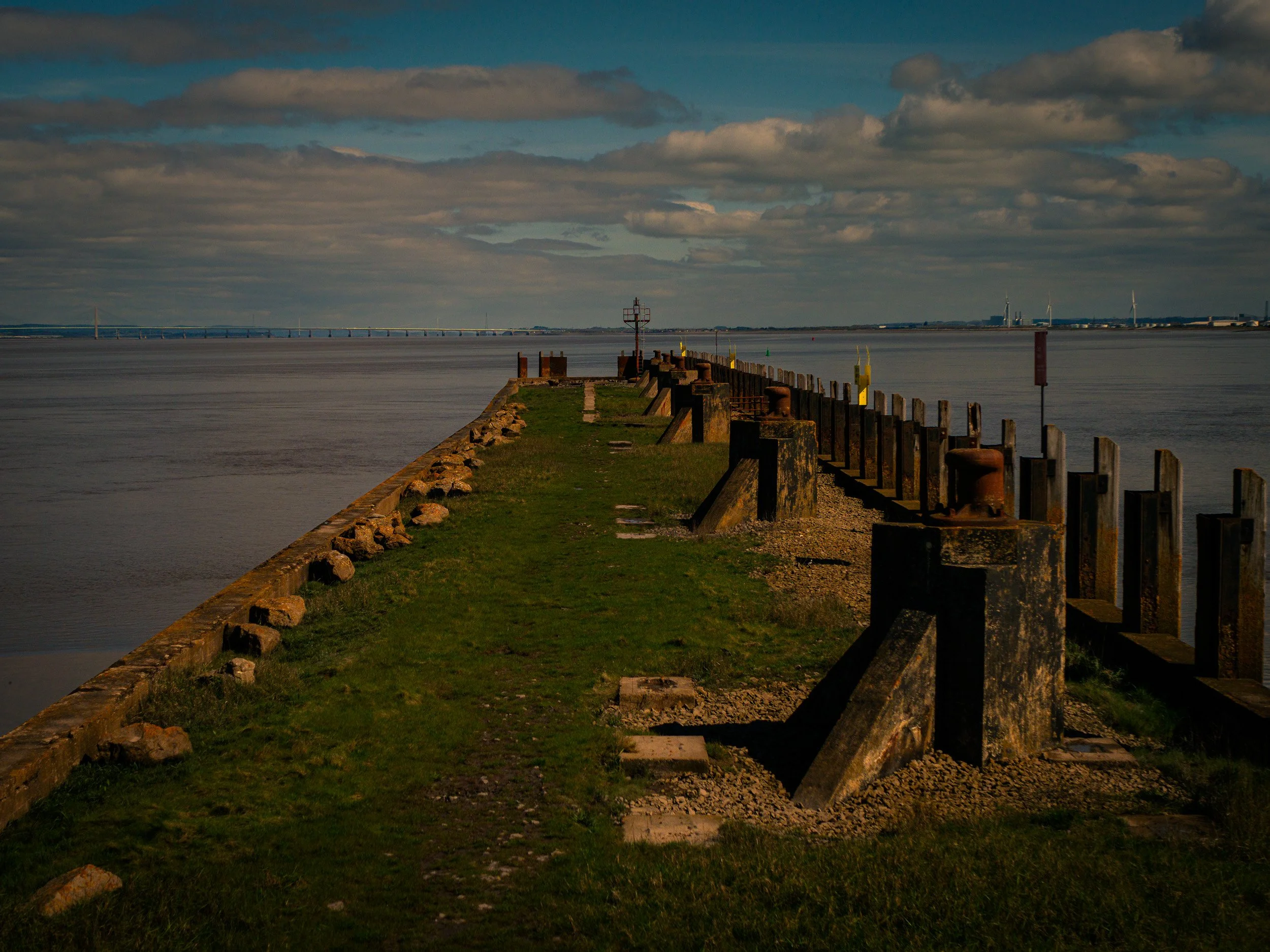A metal pier extending out into a body of water under a cloudy sky with wind turbines visible on the horizon.