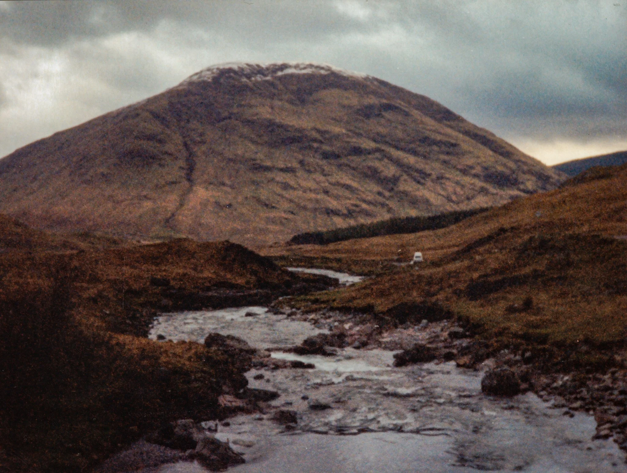 A mountain with a rocky brown surface and a snow patch at the top, under a cloudy sky, with a small river flowing through the foreground.