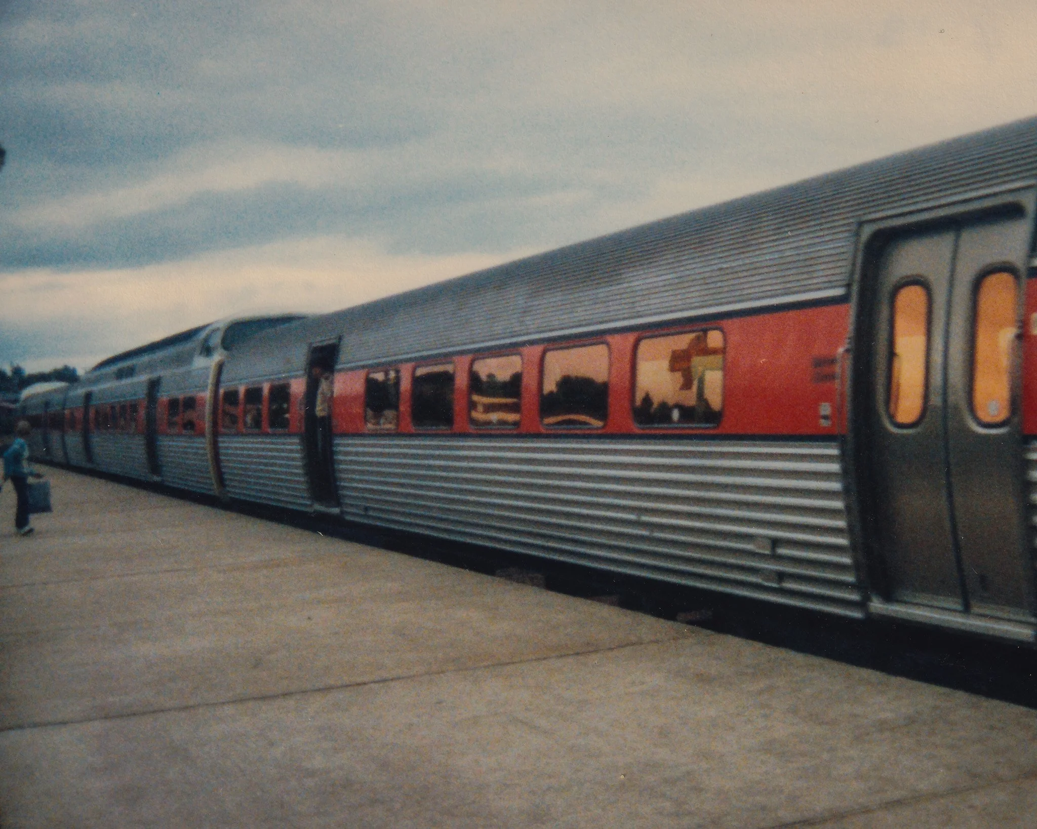 A red train with silver siding and large windows parked at a station platform, with a cloudy sky above.