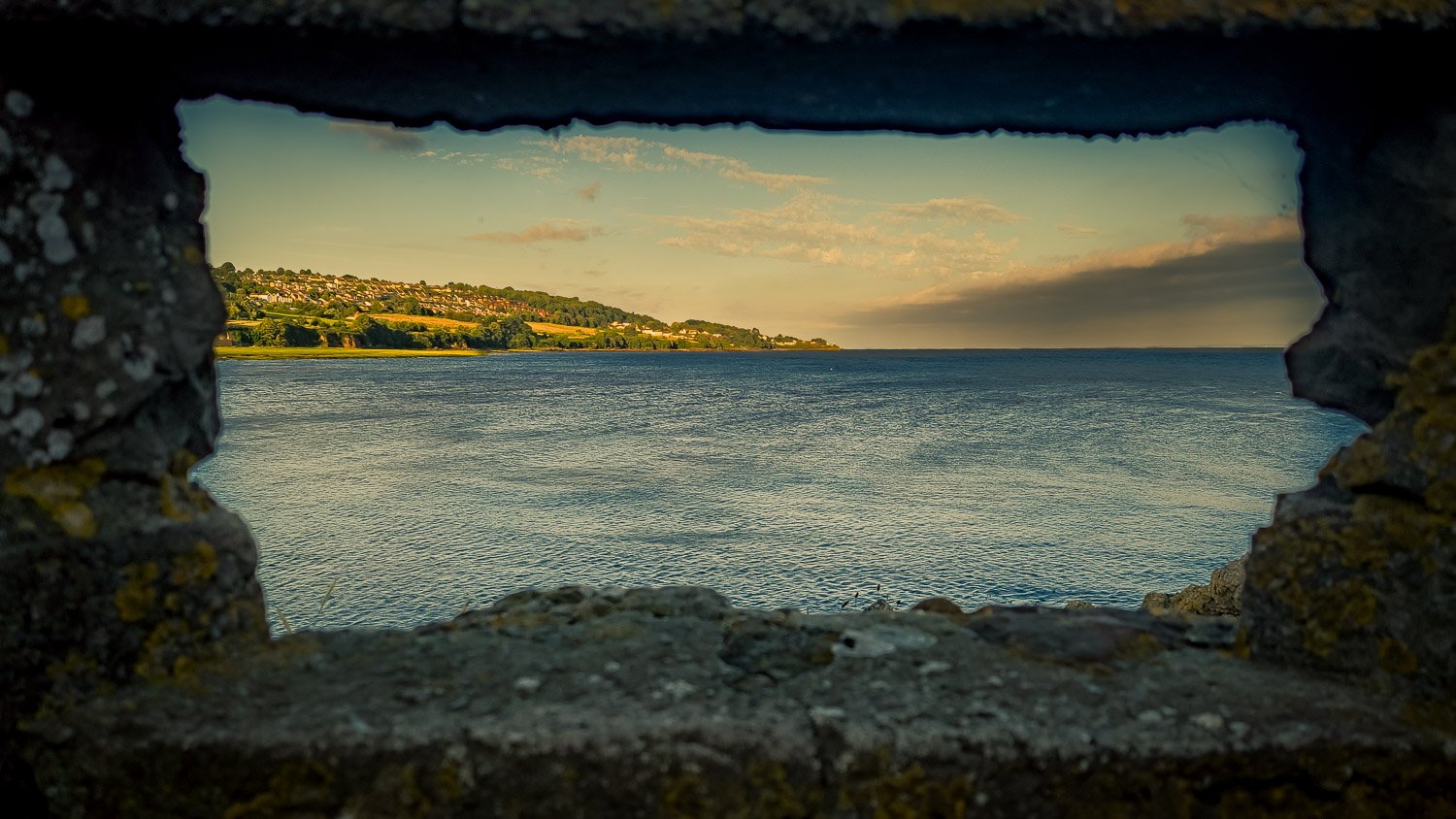 View through a rectangular hole in a stone wall showing a body of water, with hills and houses on the distant shore, under a partly cloudy sky.