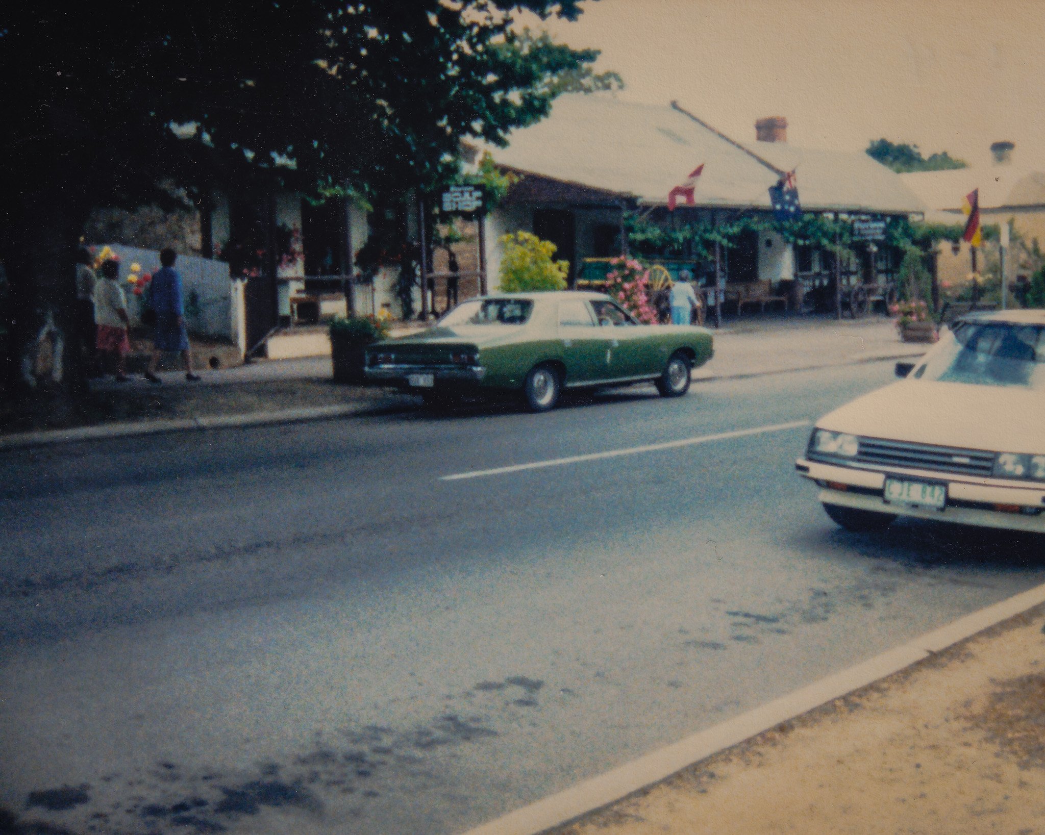 A street scene with a green and white classic car parked on the curb, and a building with a porch decorated with flags and bunting, possibly a shop or cafe, with people standing outside and a large tree nearby.