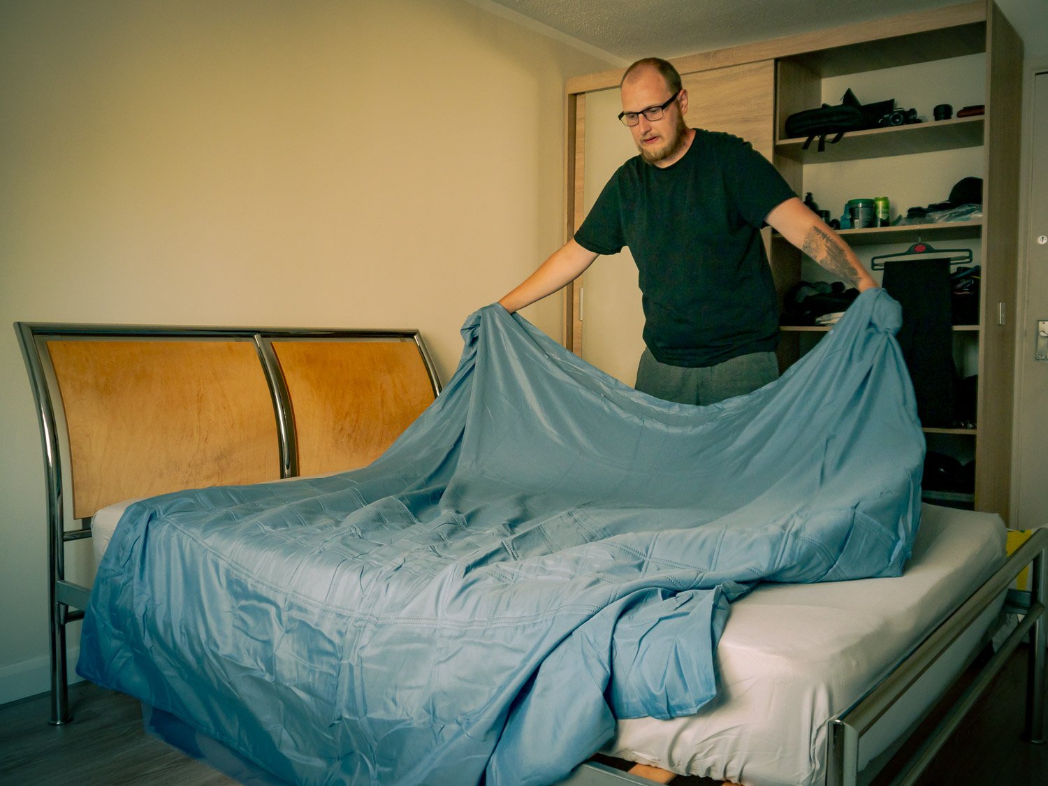 A man making a bed, pulling up a light blue bedsheet in a bedroom with wooden and metal bed frame.