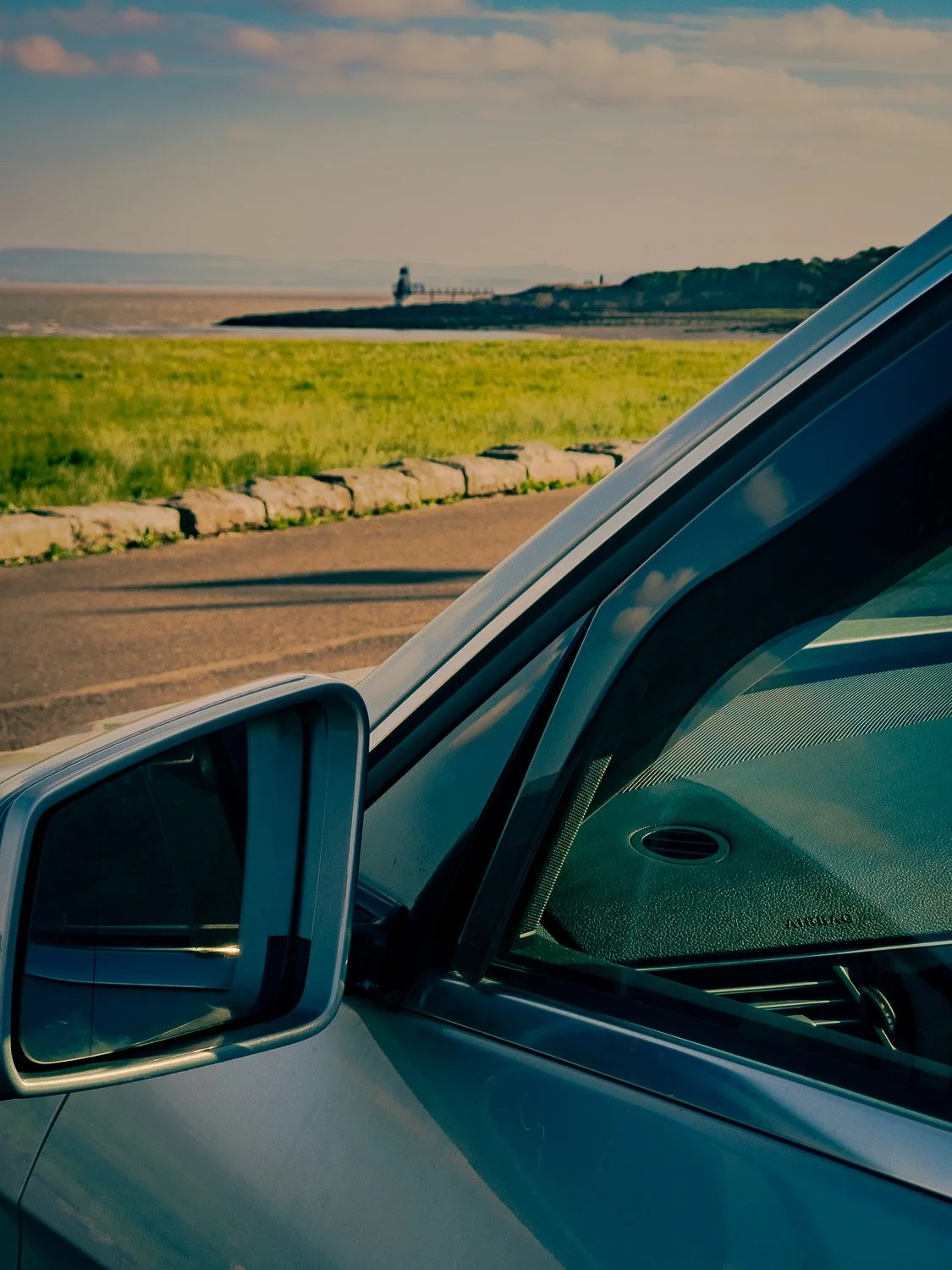 View of a coastal landscape with a grassy area, a stone barrier, and a lighthouse in the distance, seen from inside a parked vehicle.