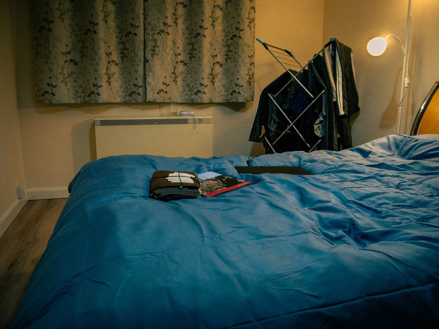 Unmade bed with a blue comforter and some papers or booklets on top. Behind the bed, a window with floral curtains, a mounted electric heater, a standing lamp, and a clothes drying rack with clothes hanging on it.