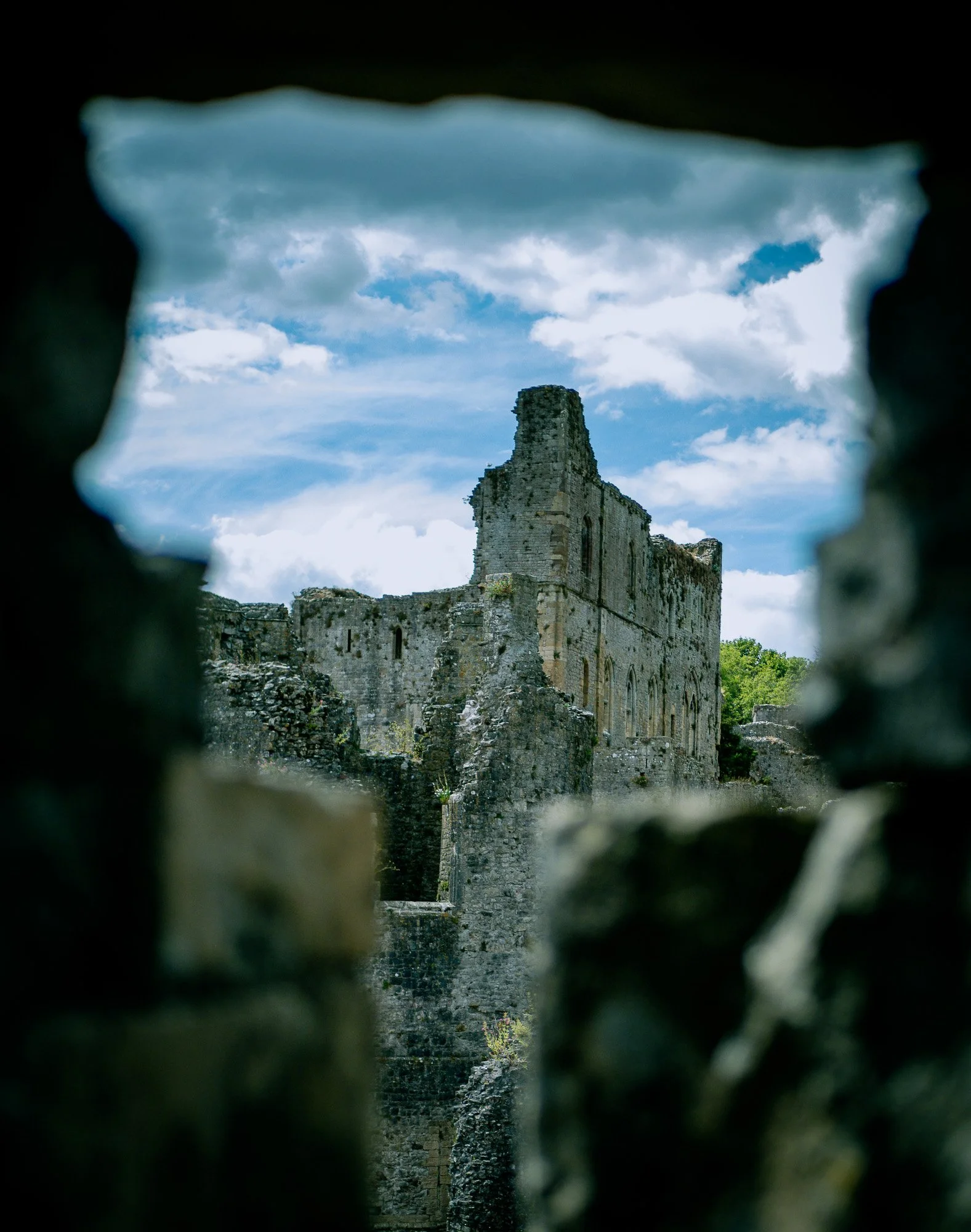 Ancient stone ruins of a castle viewed through a hole or opening in a wall, with a cloudy sky in the background.