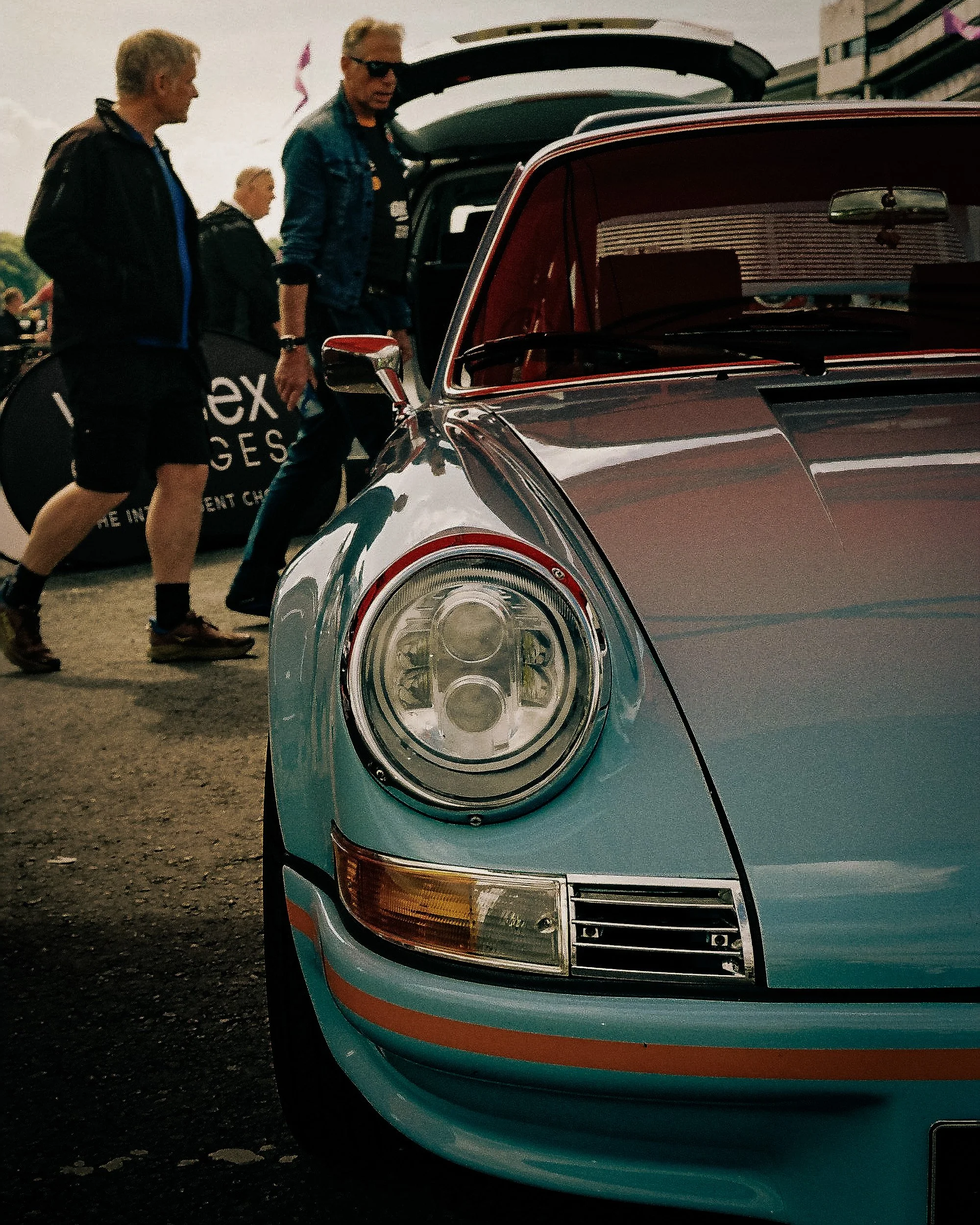 Three people near a vintage silver car with a red interior, open trunk, and distinctive headlight design at an outdoor event.