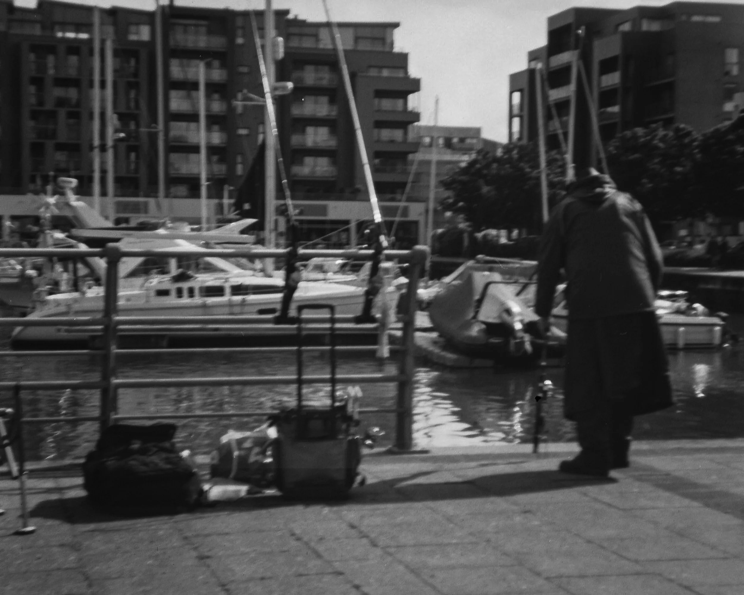 A marina with boats docked in the water, tall residential buildings in the background, a person in a hoodie walking along the waterfront, and various fences and structures near the water's edge, all captured in black-and-white.