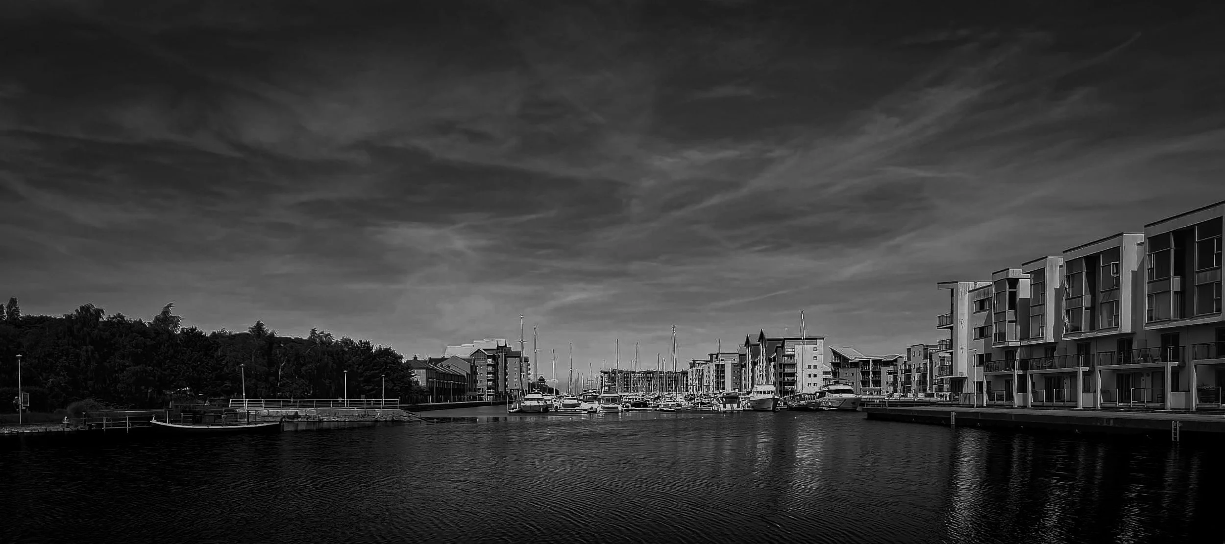 Black and white photo of a marina with boats docked, modern apartment buildings along the waterfront, and a cloudy sky.