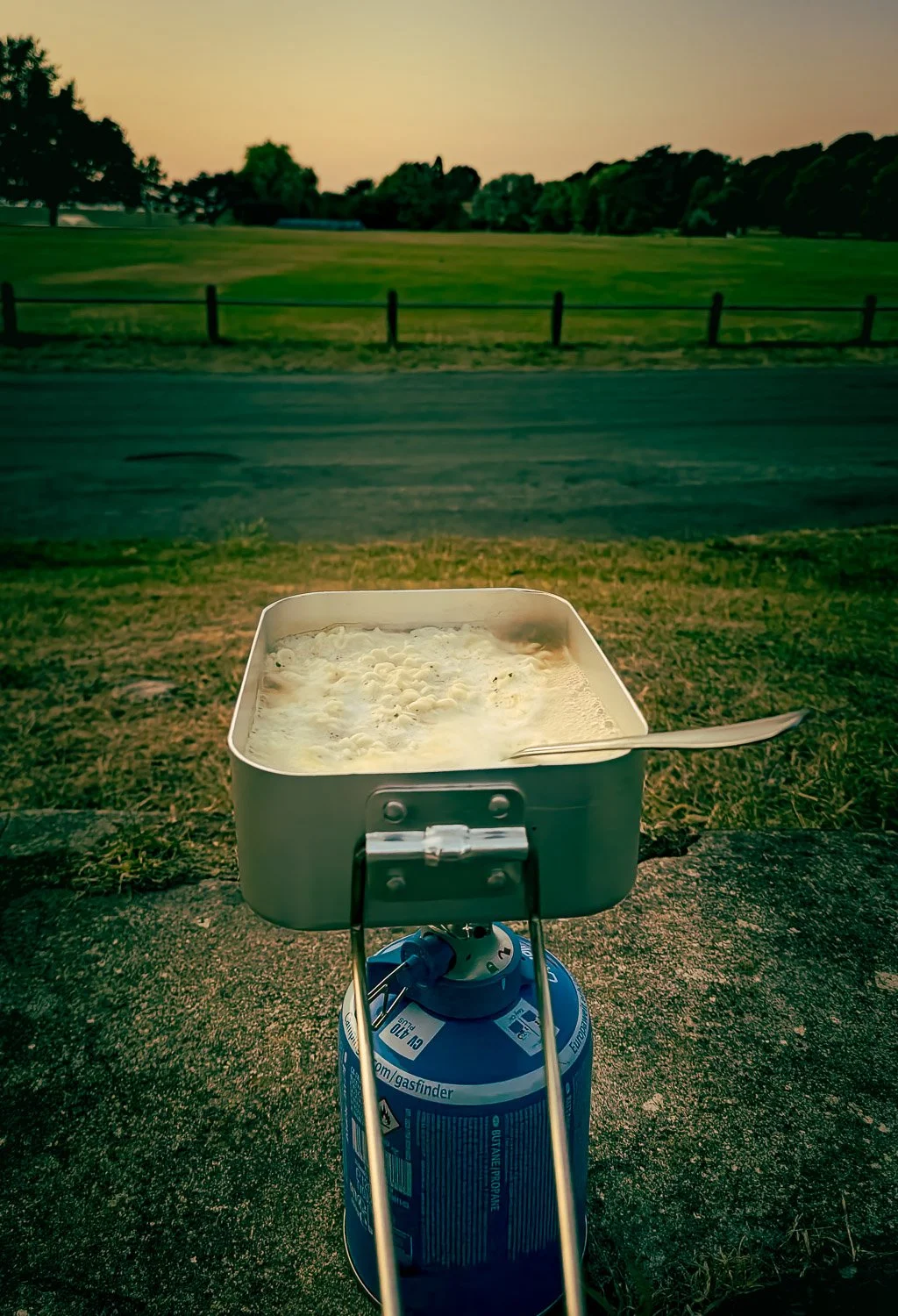 A small portable stove with a pot of boiling water and food on top, outdoors during sunset with green fields and trees in the background.