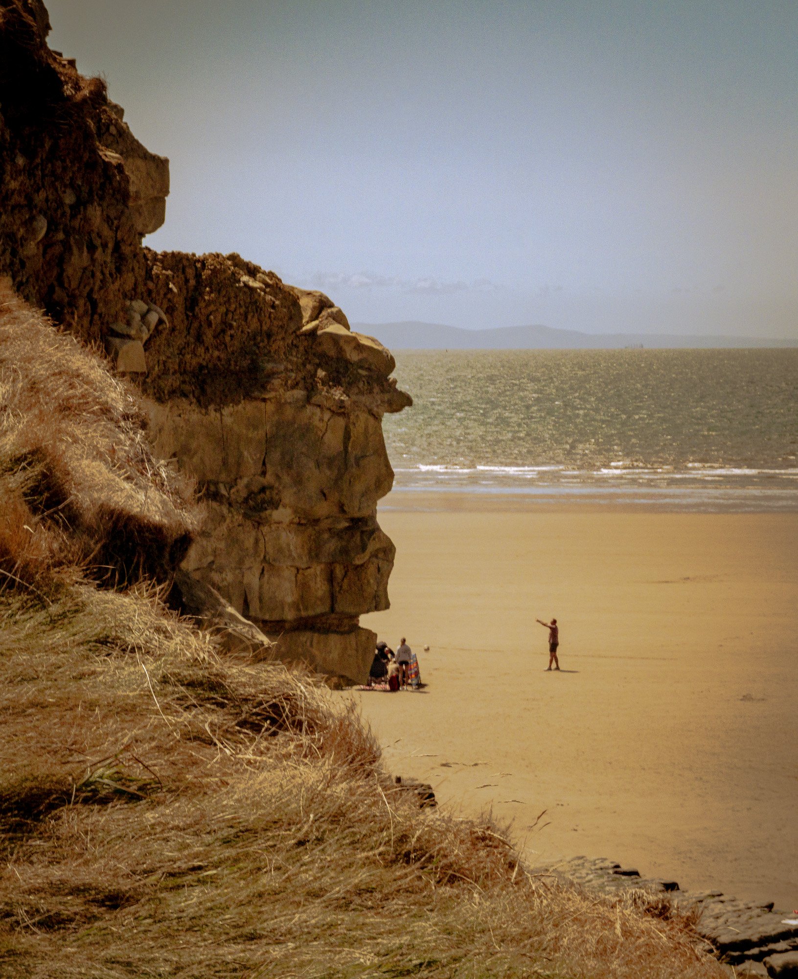 People on a sandy beach near a large rocky cliff with dry grass, water, and mountains in the background.