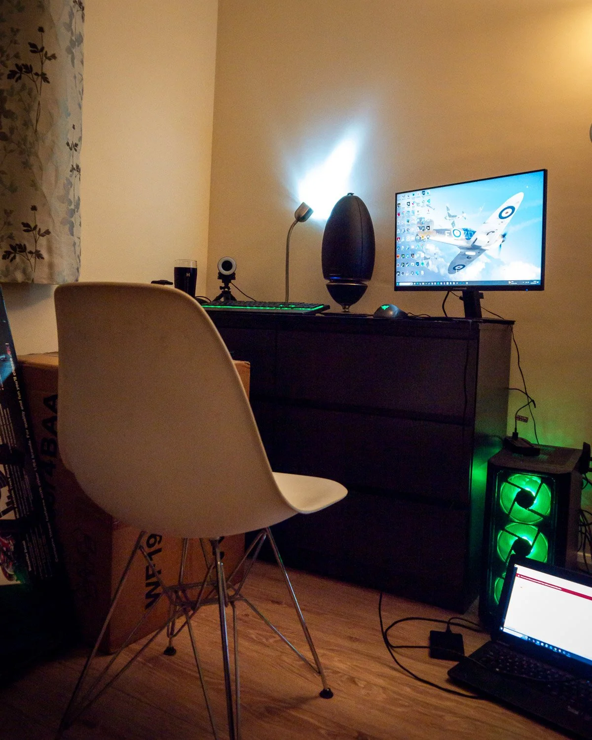 A computer desk with a monitor displaying a plane in the sky, a keyboard, a webcam, a speaker, and a lamp. There's a white chair in front of the desk, a box under the chair, and a gaming PC with green lights on the right. A laptop is on the floor con