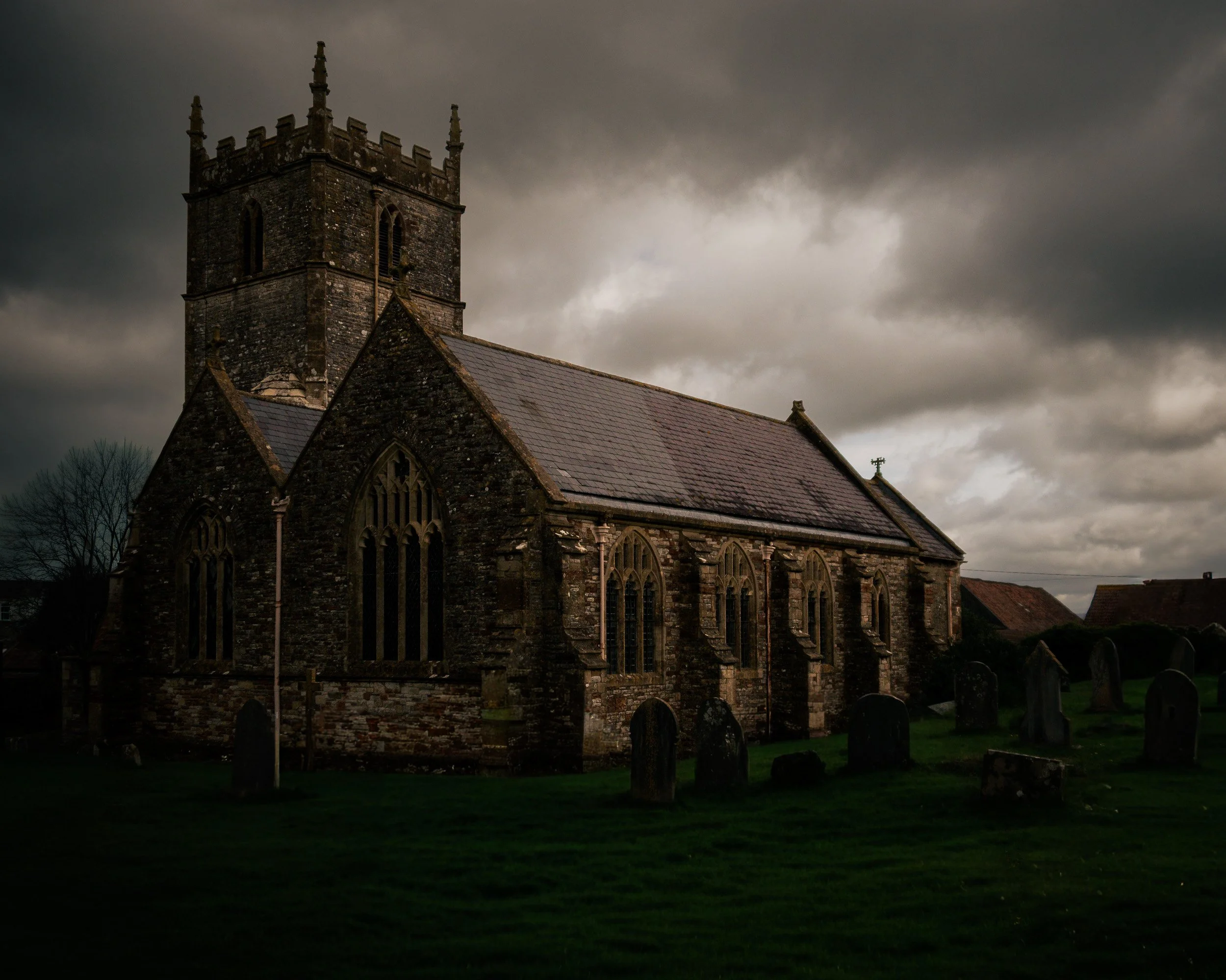 An old stone church with gothic windows and a steeple, under a cloudy, dark sky, surrounded by a graveyard with gravestones and a grassy area.