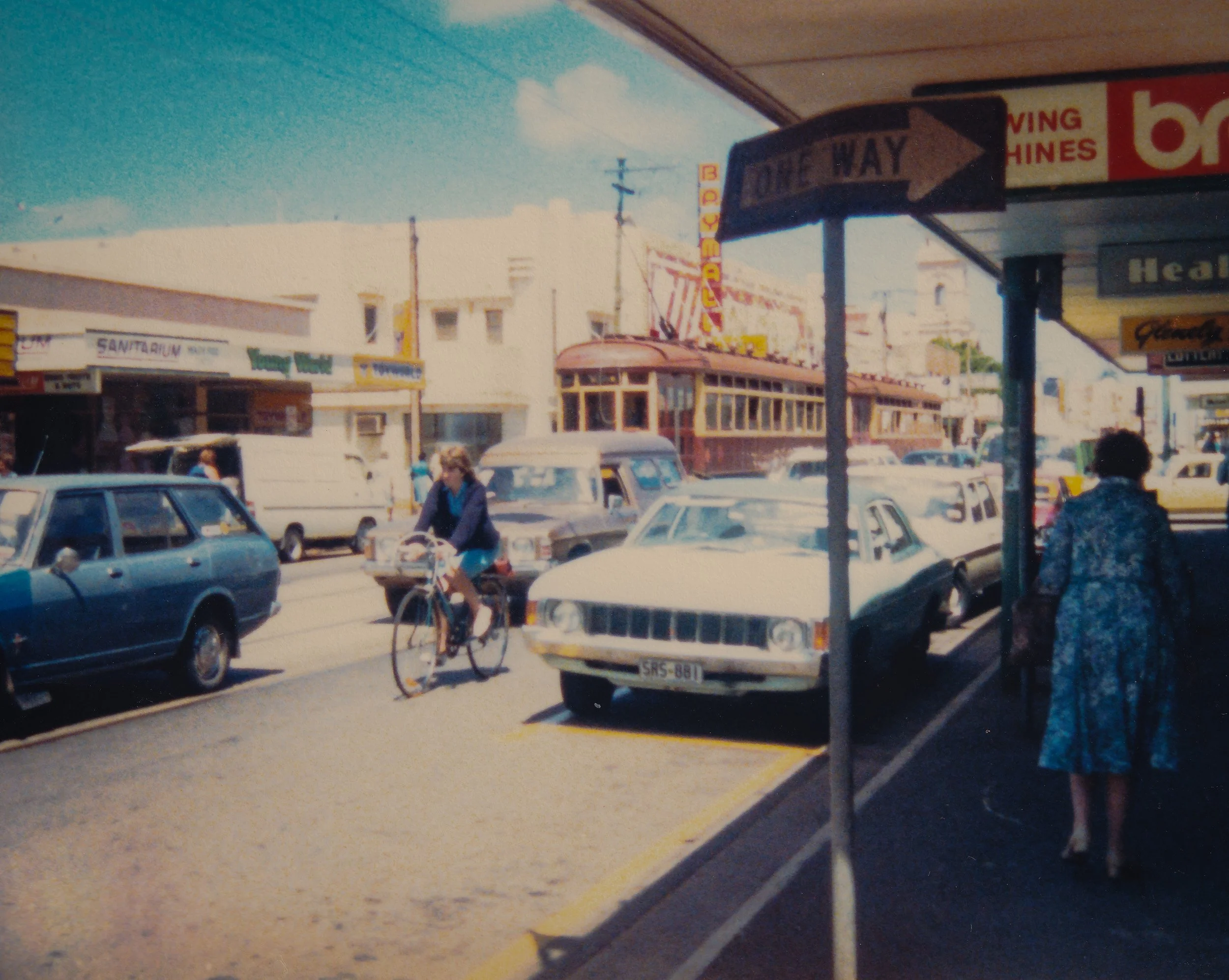 A street scene with cars parked along the street, pedestrians walking, a cyclist in motion, signs for businesses, and a clear blue sky.