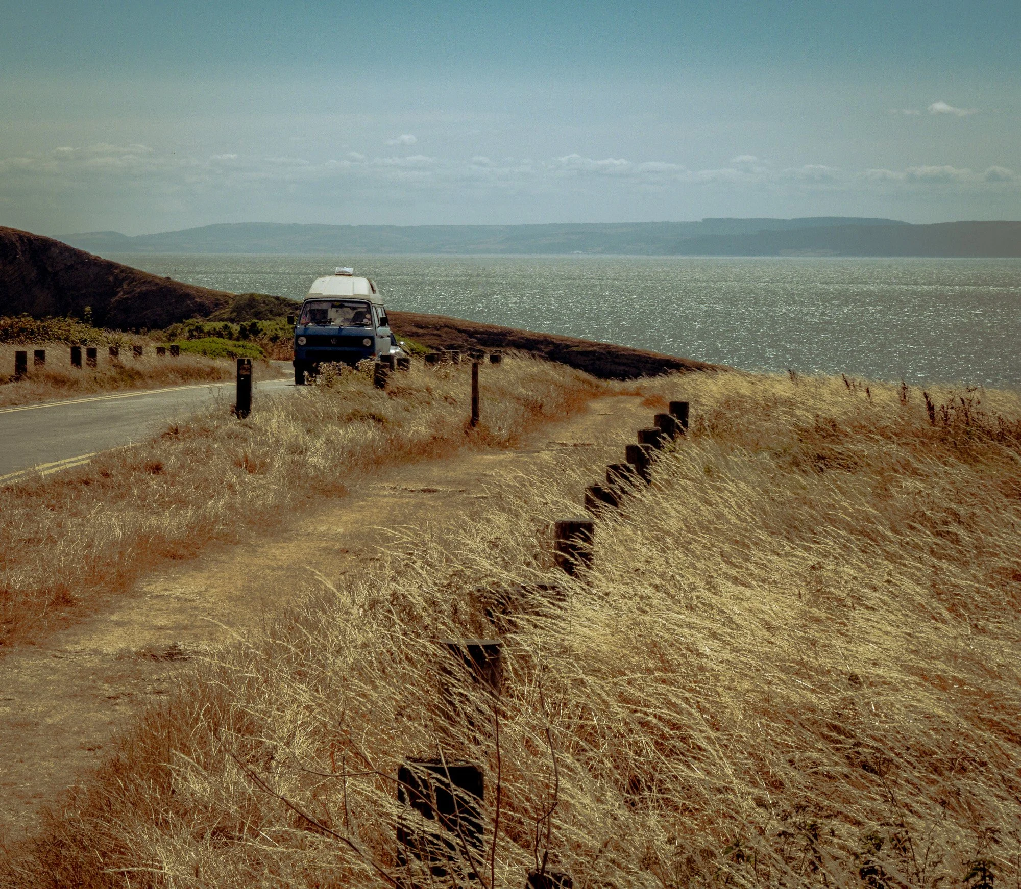 A winding road along a grassy hillside overlooking a large body of water with distant hills on the horizon under a blue sky with scattered clouds.