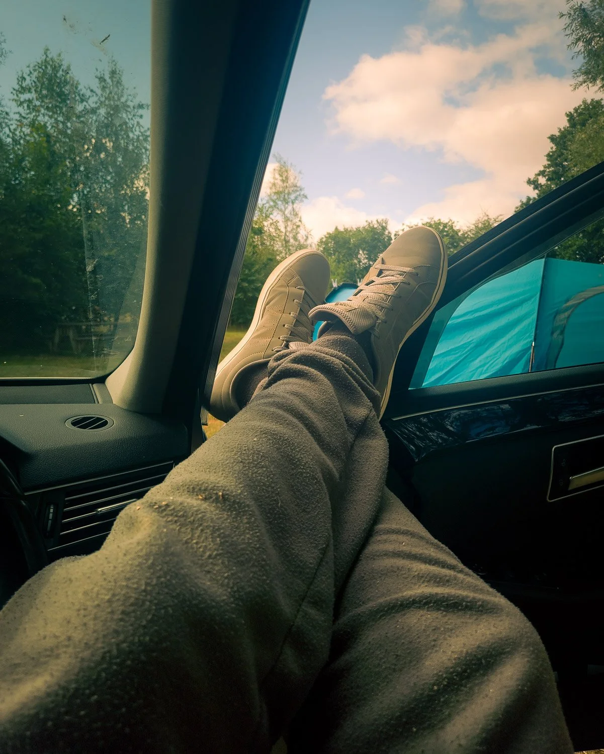 Person relaxing with legs up on dashboard of car, window view of trees and cloudy sky outside