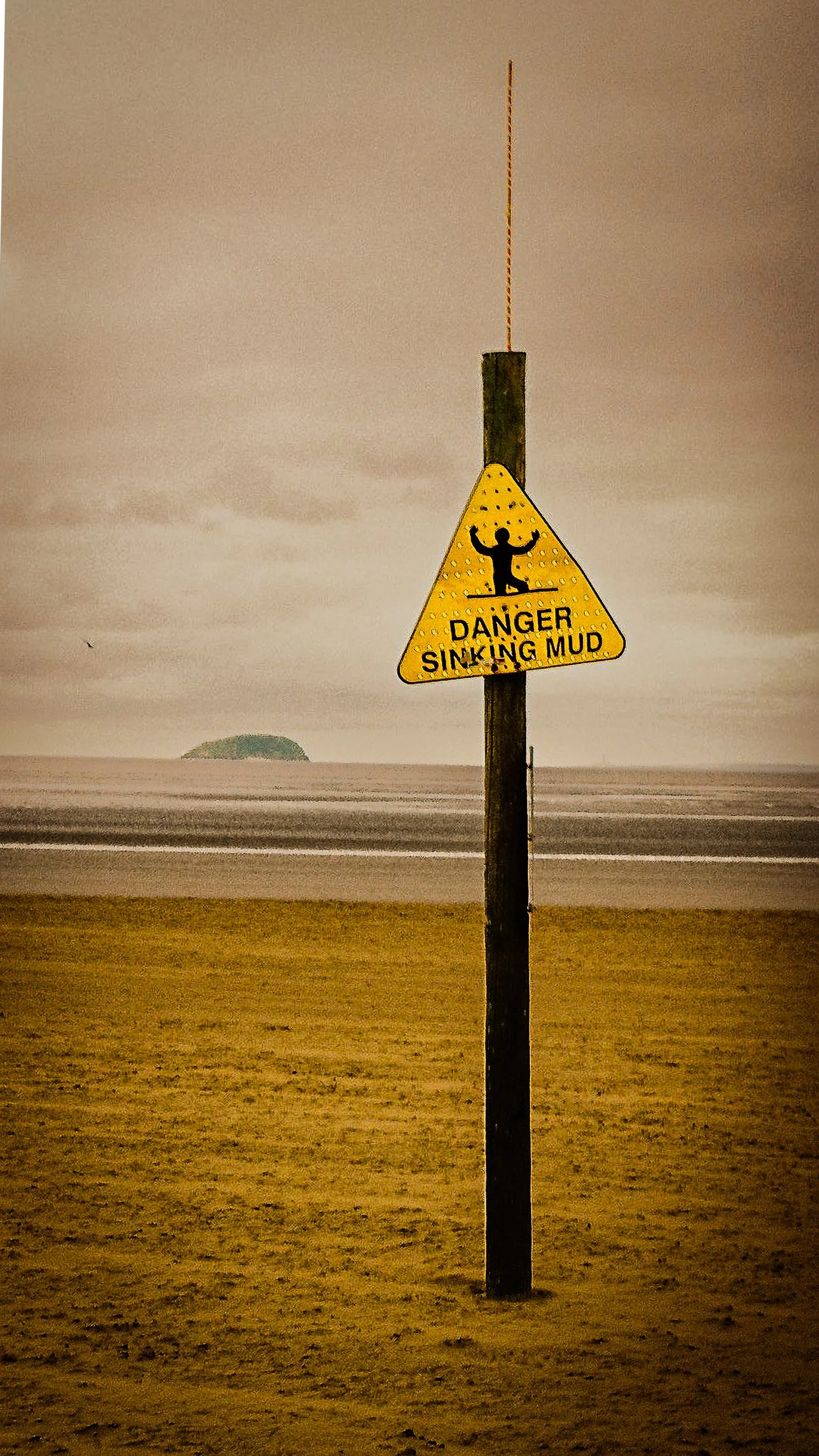 A yellow warning sign with a silhouette of a person falling, reading 'Danger Sinking Mud,' and a small island in the background over a sandy beach.