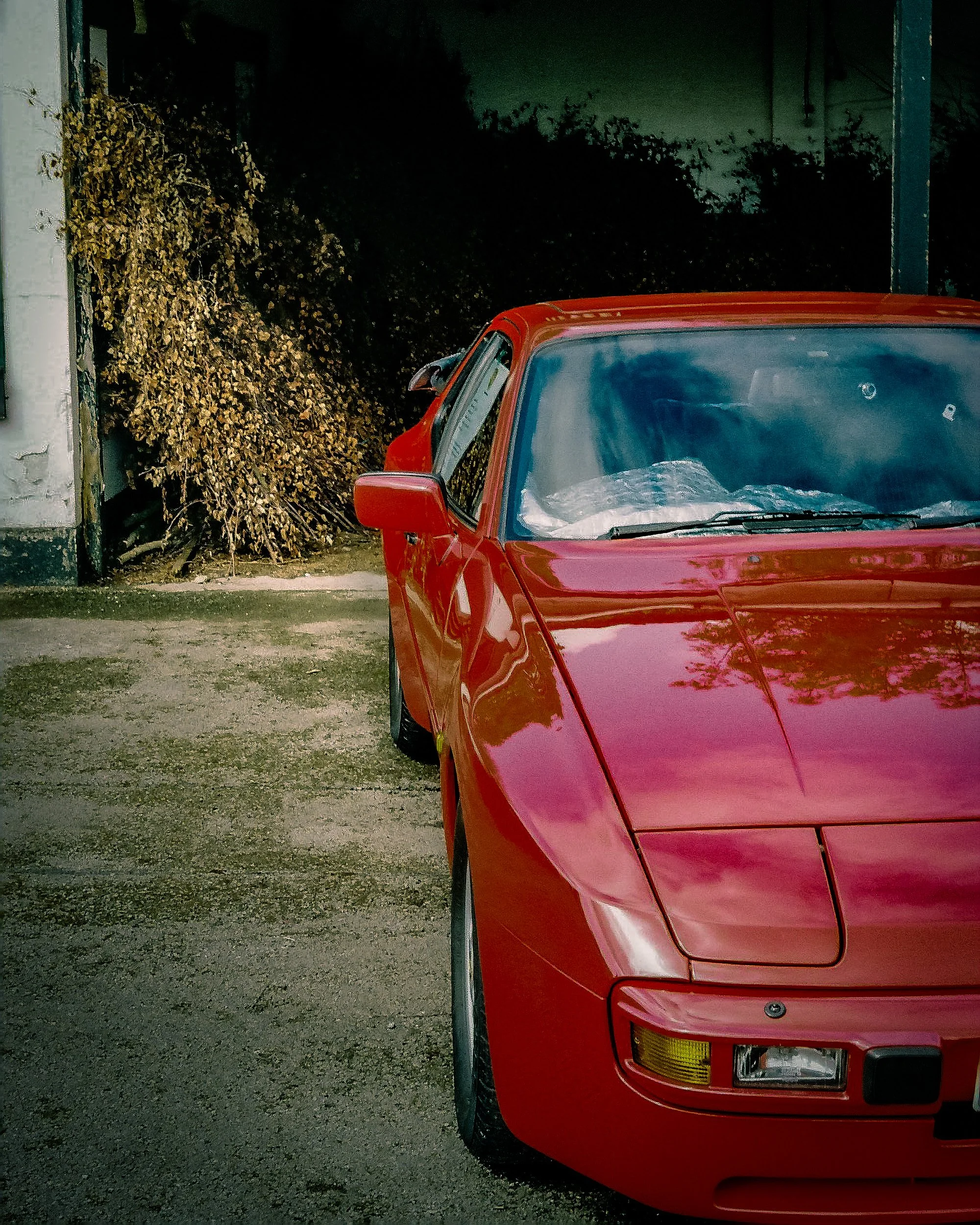 Front of a red sports car parked on a gravel surface with foliage and a building in the background during nighttime.