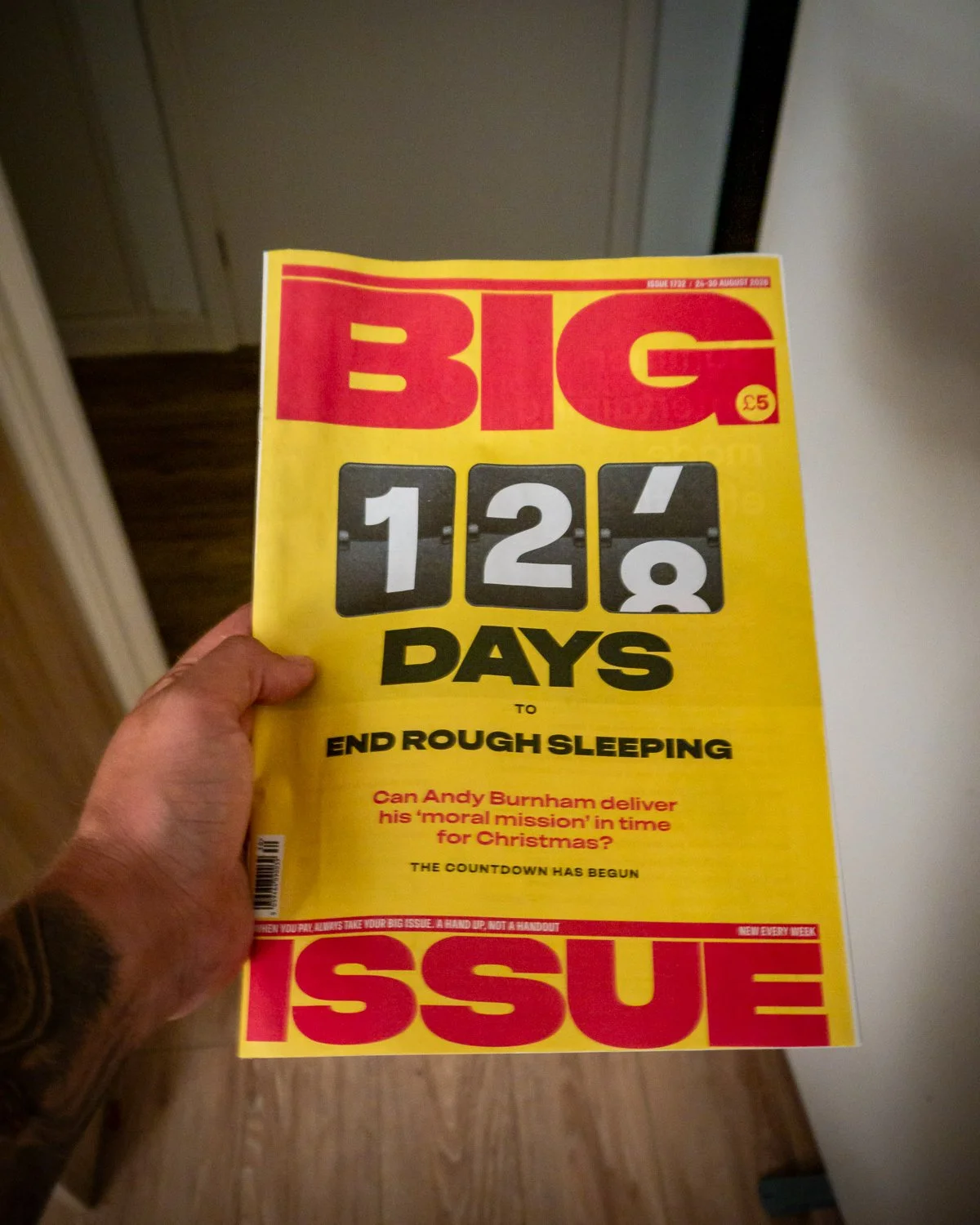 A person holding a magazine titled "BIG ISSUE" with a yellow background, large red title, and a countdown of 124 days before ending rough sleeping. The magazine features information about Andy Burnham and a note about another issue.